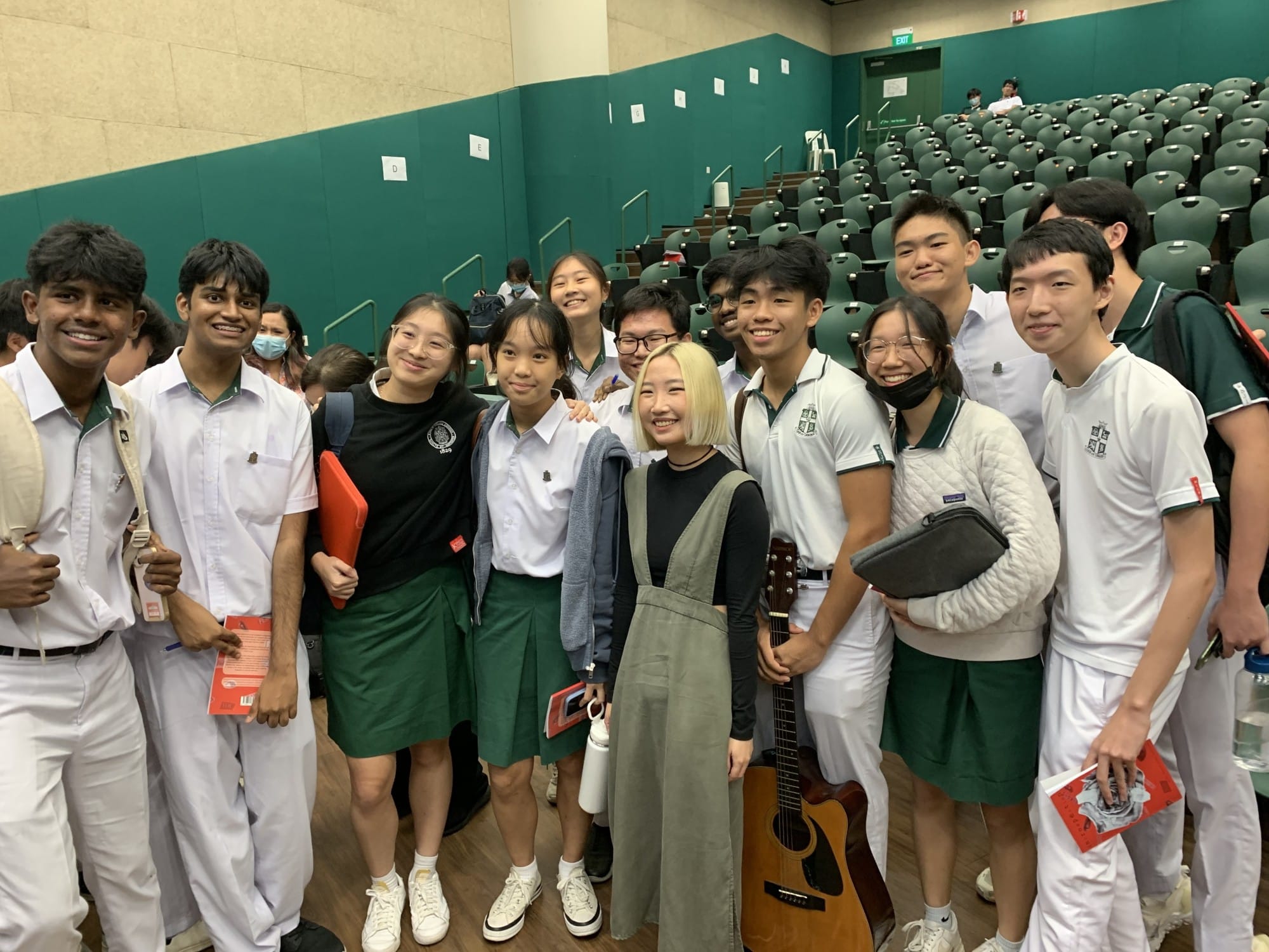 A group of smiling school students in green and white uniforms pose with a young woman holding a guitar in an auditorium.