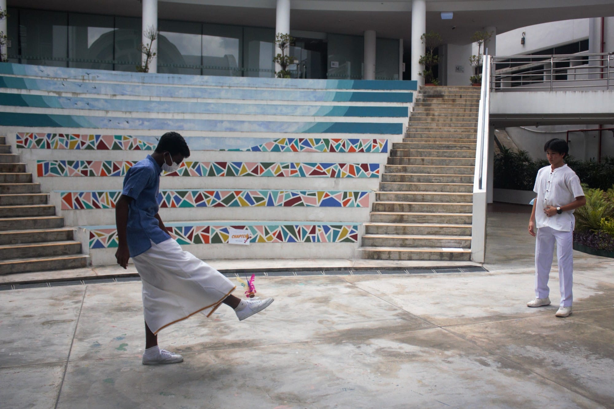 Two boys in white clothing stand near colorful mosaic-decorated stairs, one kicking a small object on the ground.