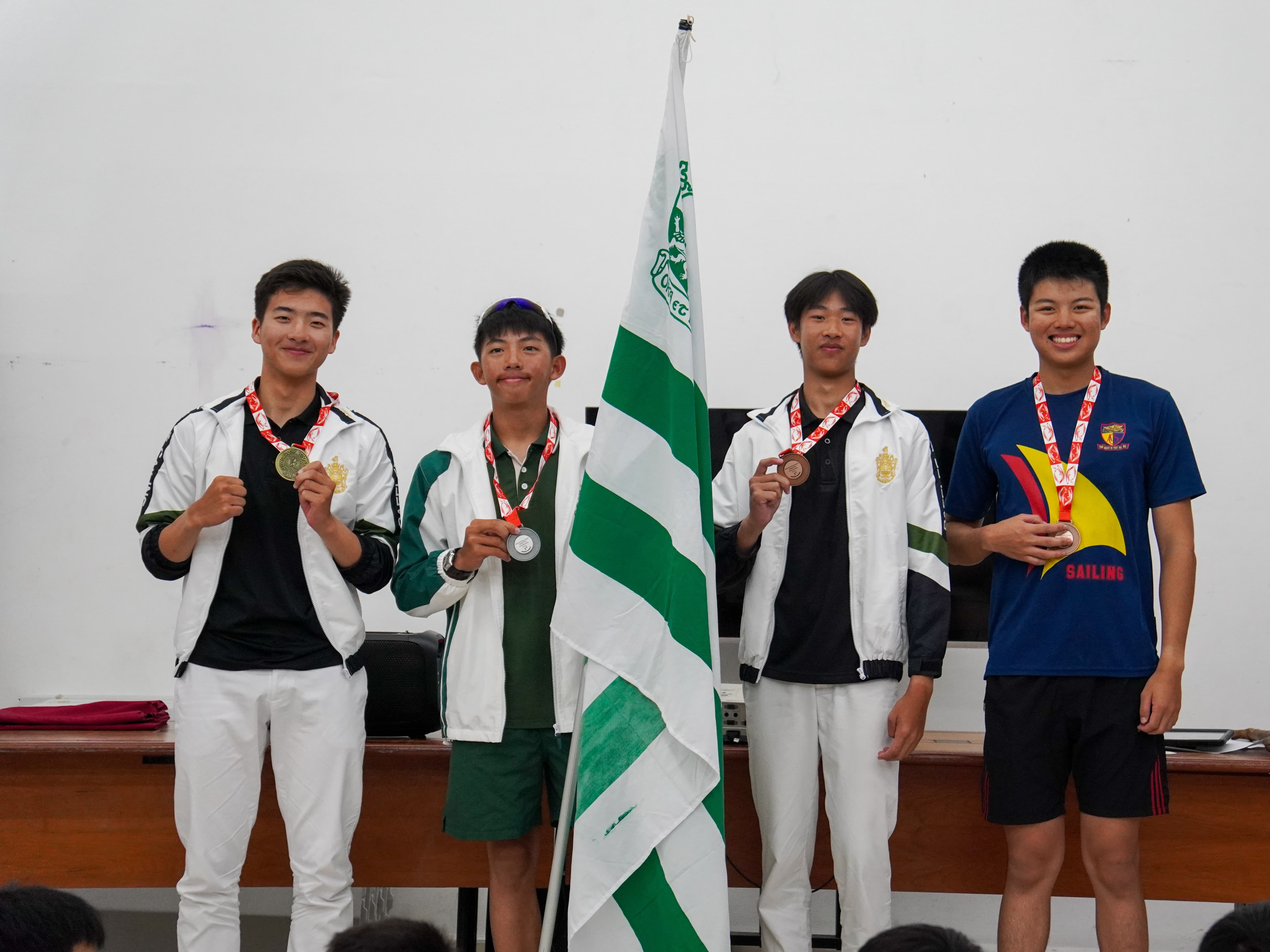 Four young male athletes smile while displaying gold, silver, and bronze medals, standing beside a green and white flag.