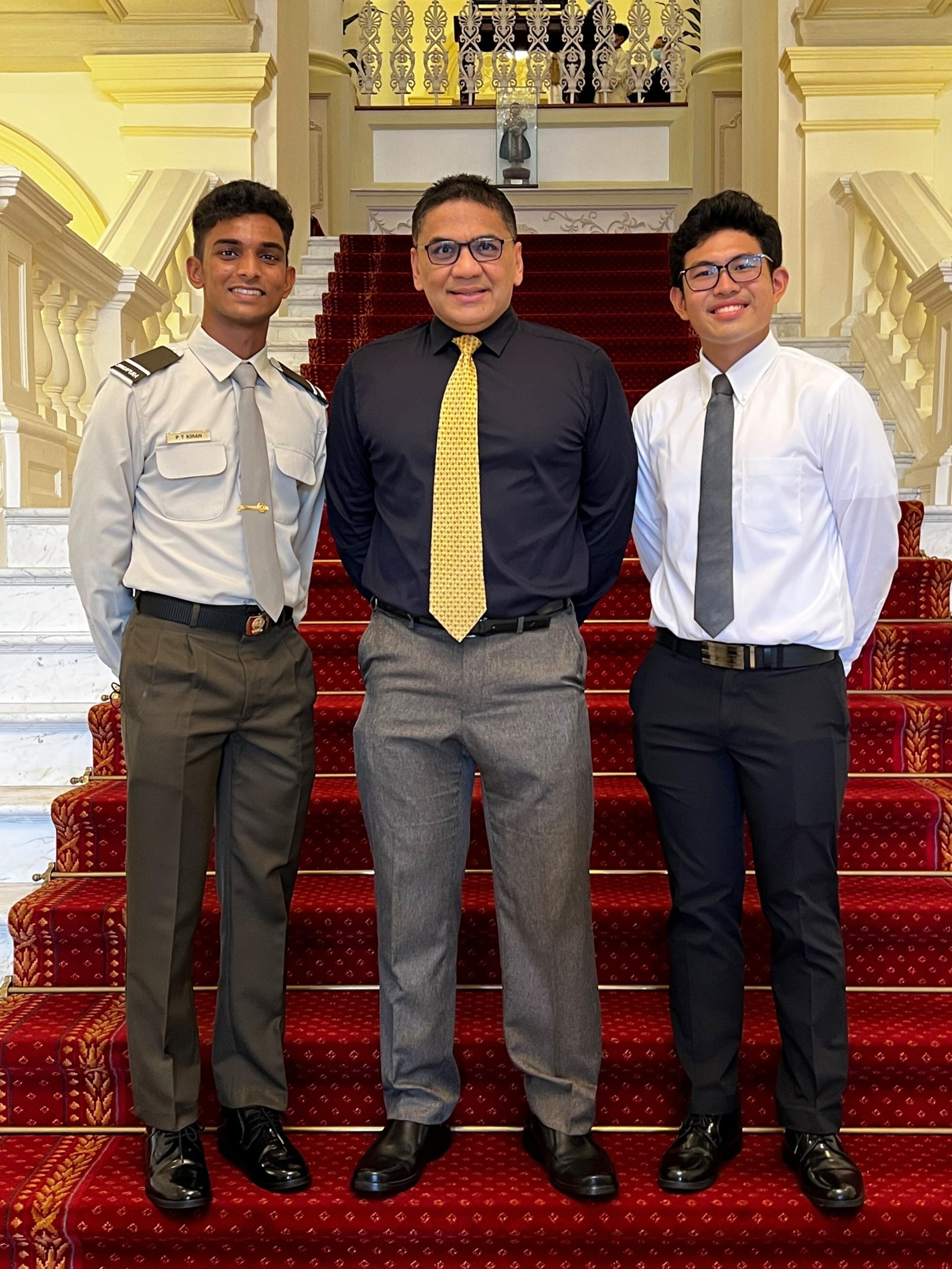 Three men in formal attire stand smiling on red-carpeted stairs in an ornate building with white columns.