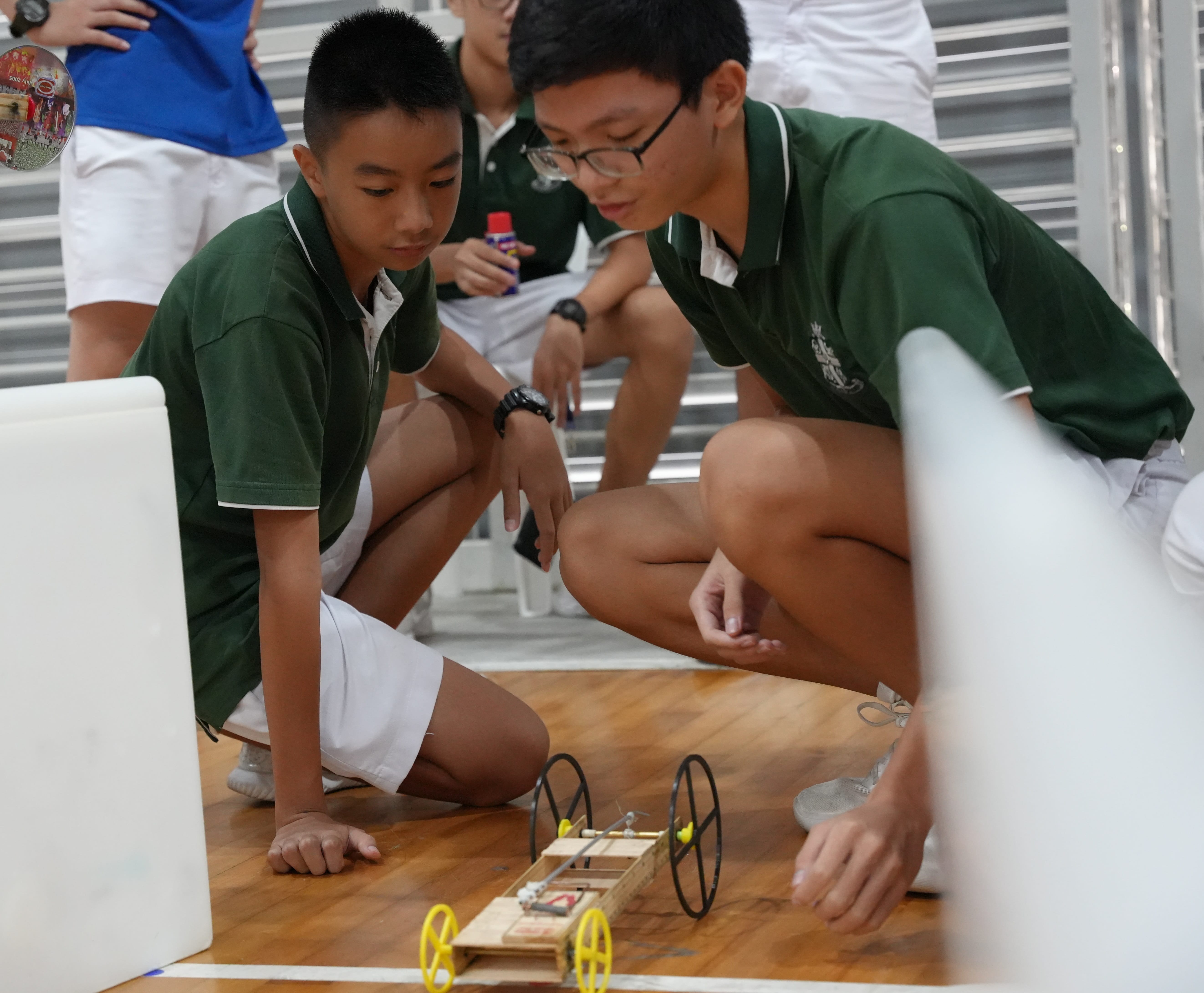 Two students in green uniforms crouch on a gym floor, watching a small wheeled mousetrap car.