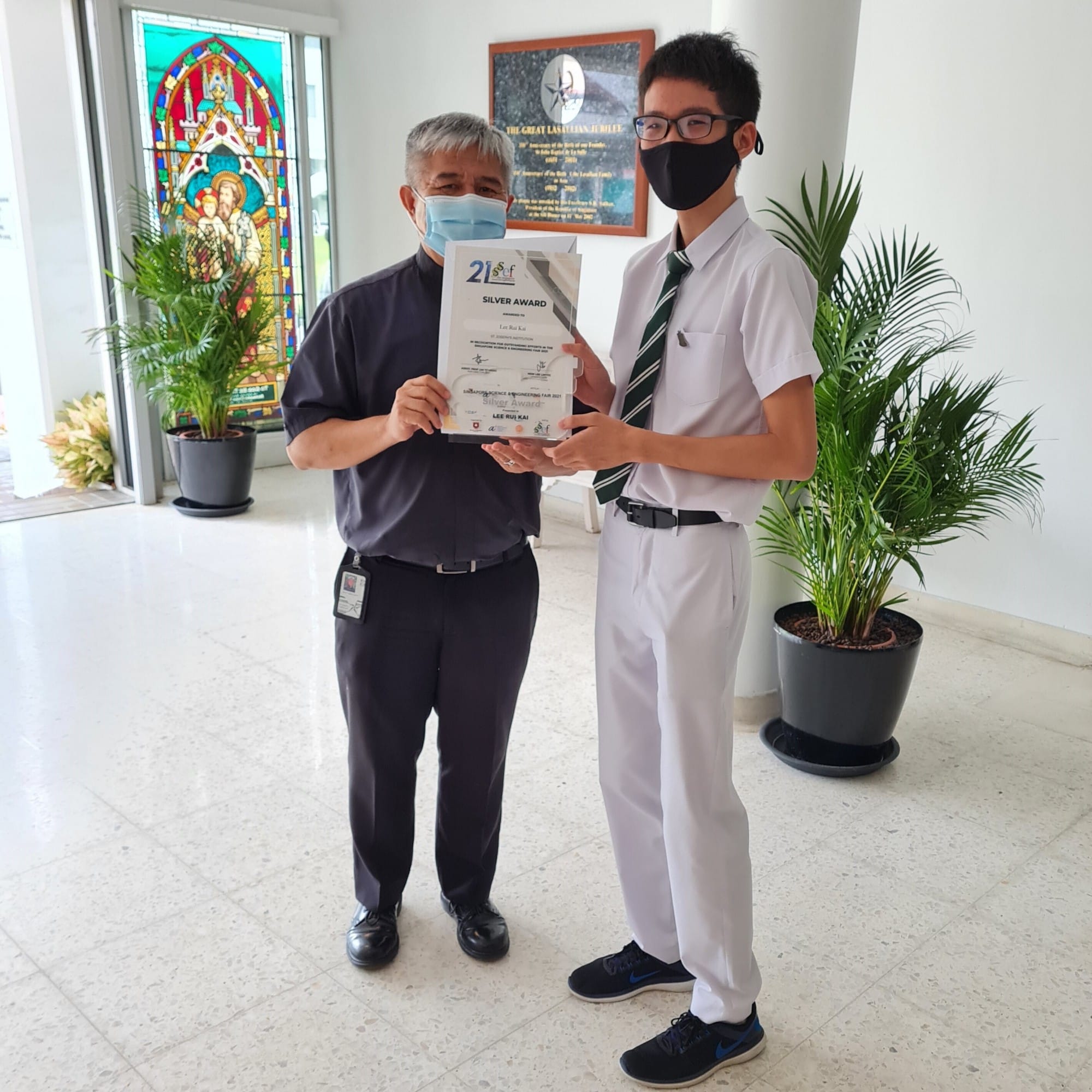 Two masked men hold a Silver Award certificate indoors near potted plants and a stained glass window.