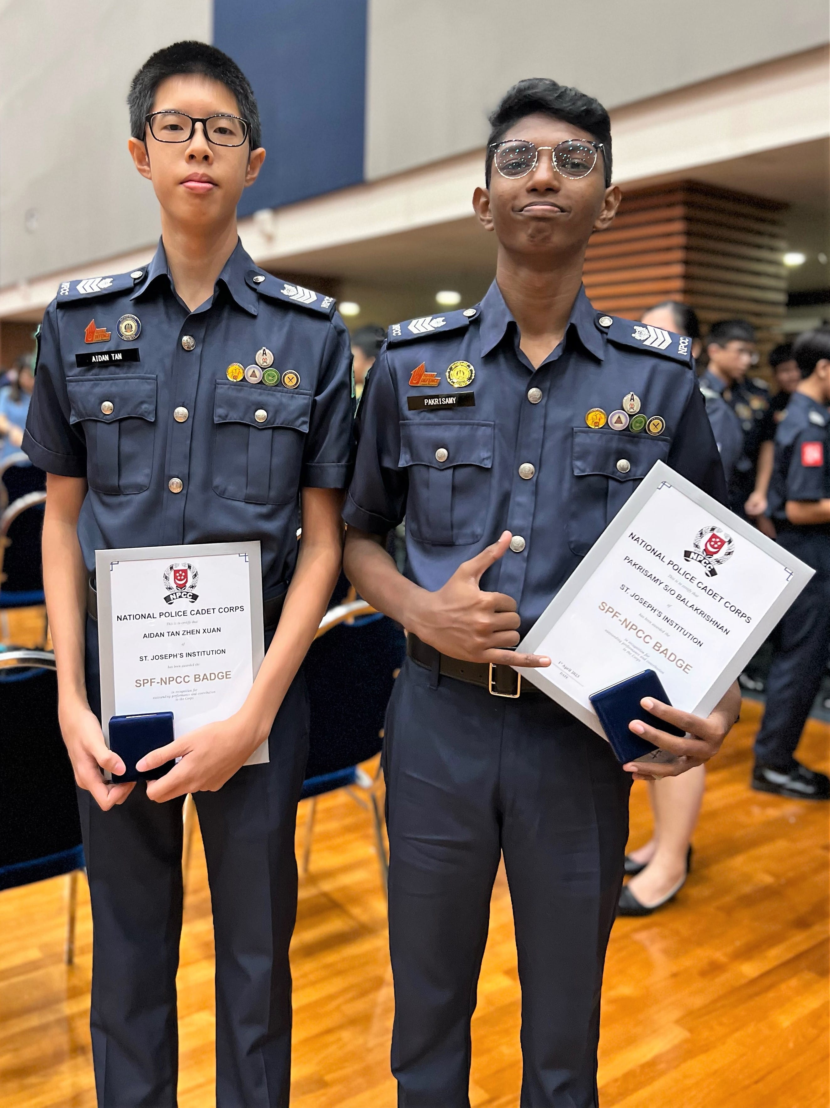 Two young cadets in Navy NPCC uniforms hold SPF-NPCC Badge certificates and medal cases indoors.