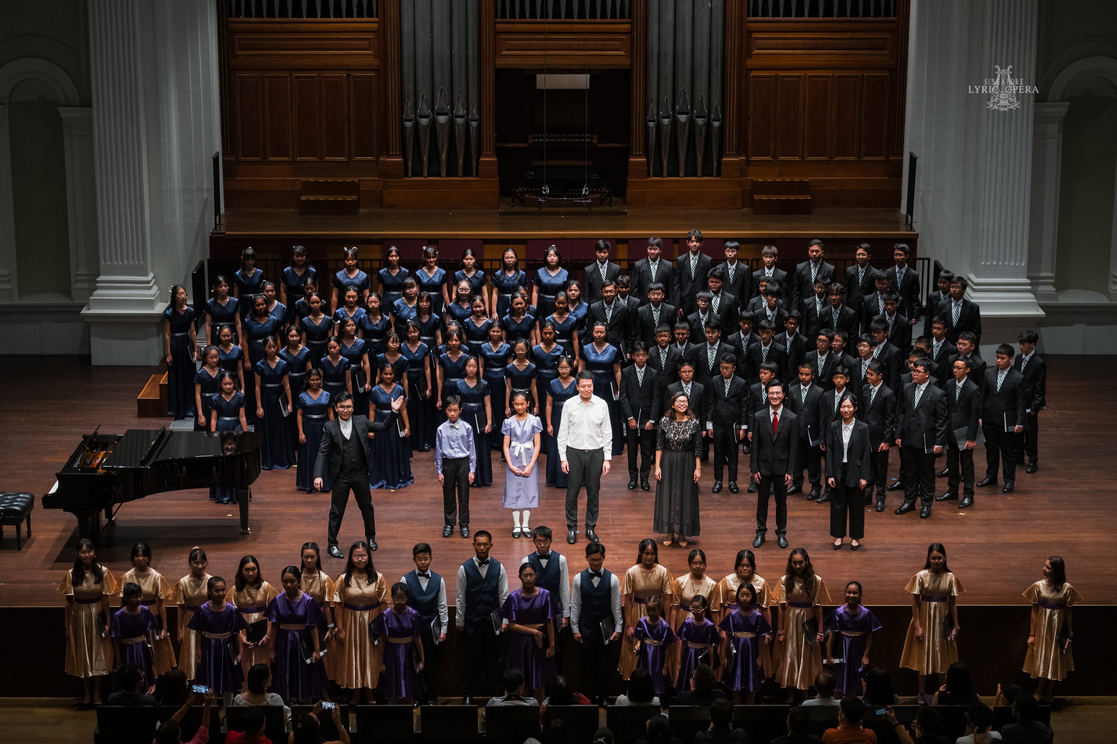 Large youth choir in uniform standing on a concert hall stage with a grand piano and pipe organ backdrop.