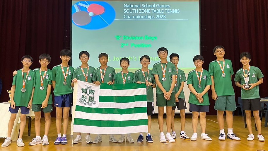 A team of students in green jerseys with silver medals pose holding a green and white striped flag, in front of a "South Zone Table Tennis Championships 2023 'B' Division Boys 2nd Position" screen.