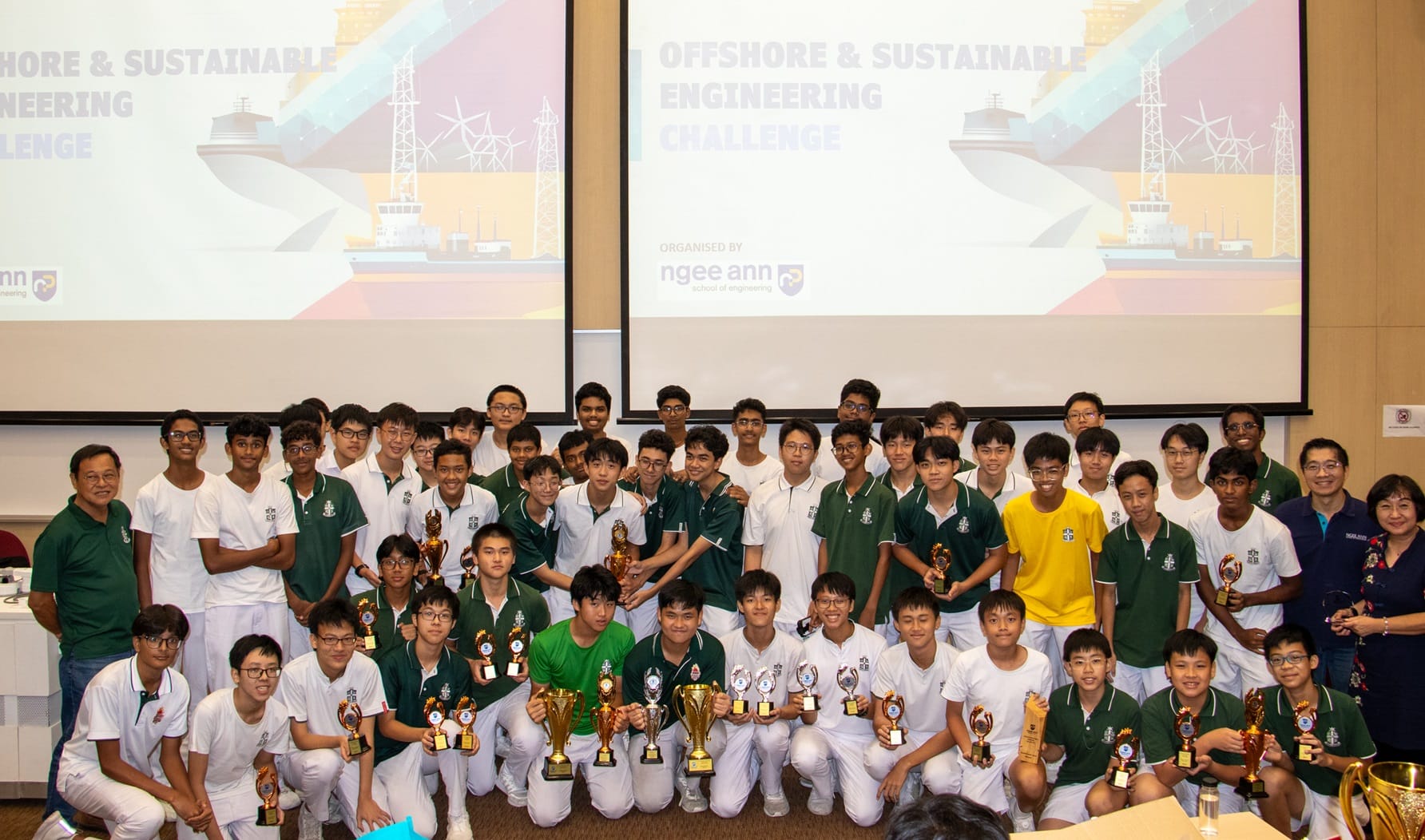 A large group of students holding trophies pose in front of screens showing Offshore and Sustainable Engineering Challenge.
