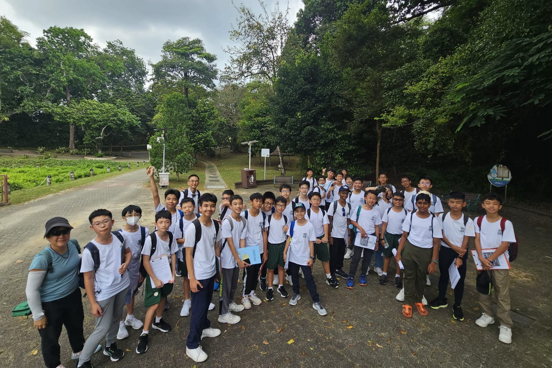 A large group of students in white shirts with backpacks pose together on a tree-lined park path with a teacher.