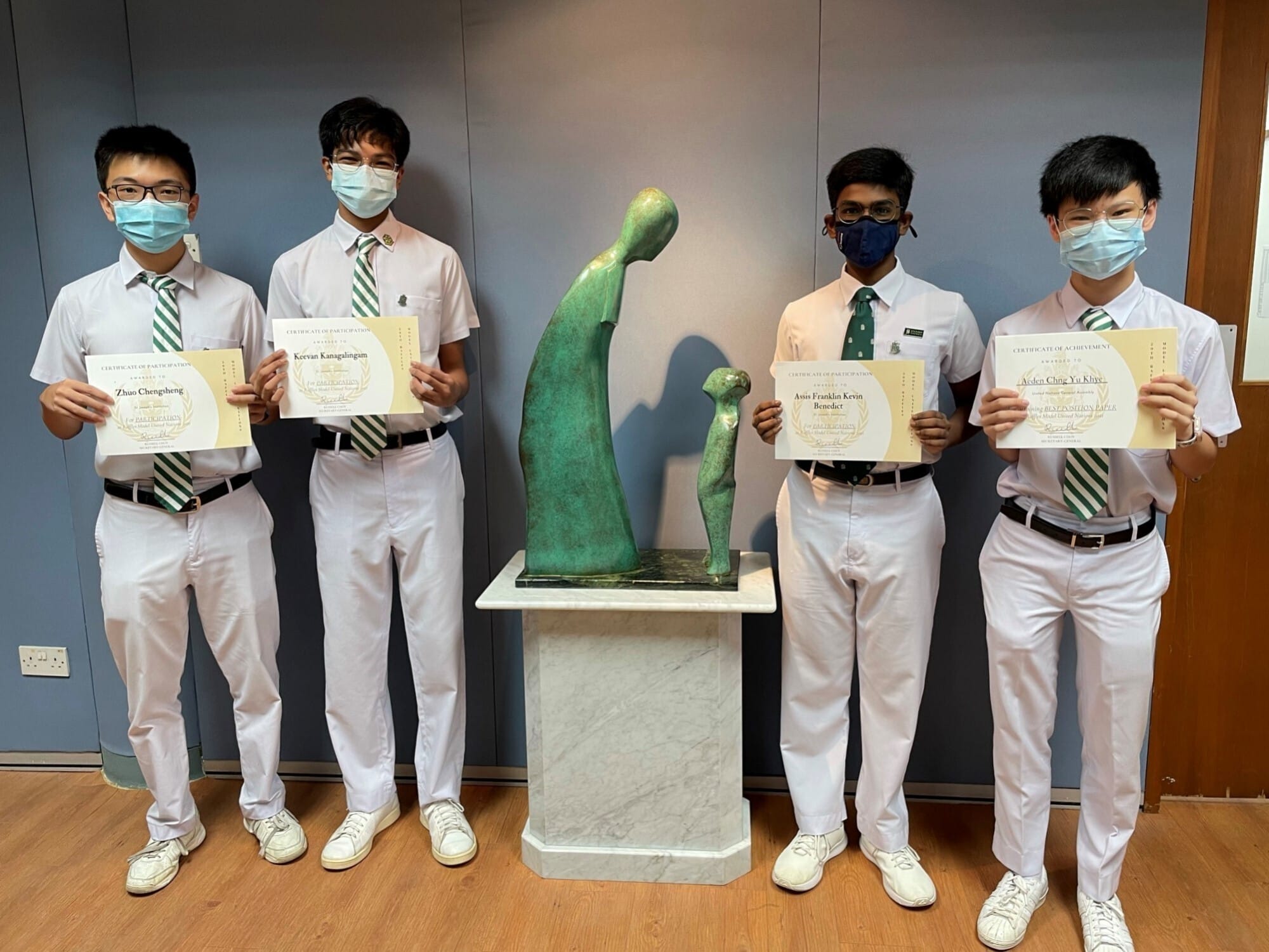 Four masked male students in white uniforms hold certificates beside a green bronze sculpture on a pedestal.