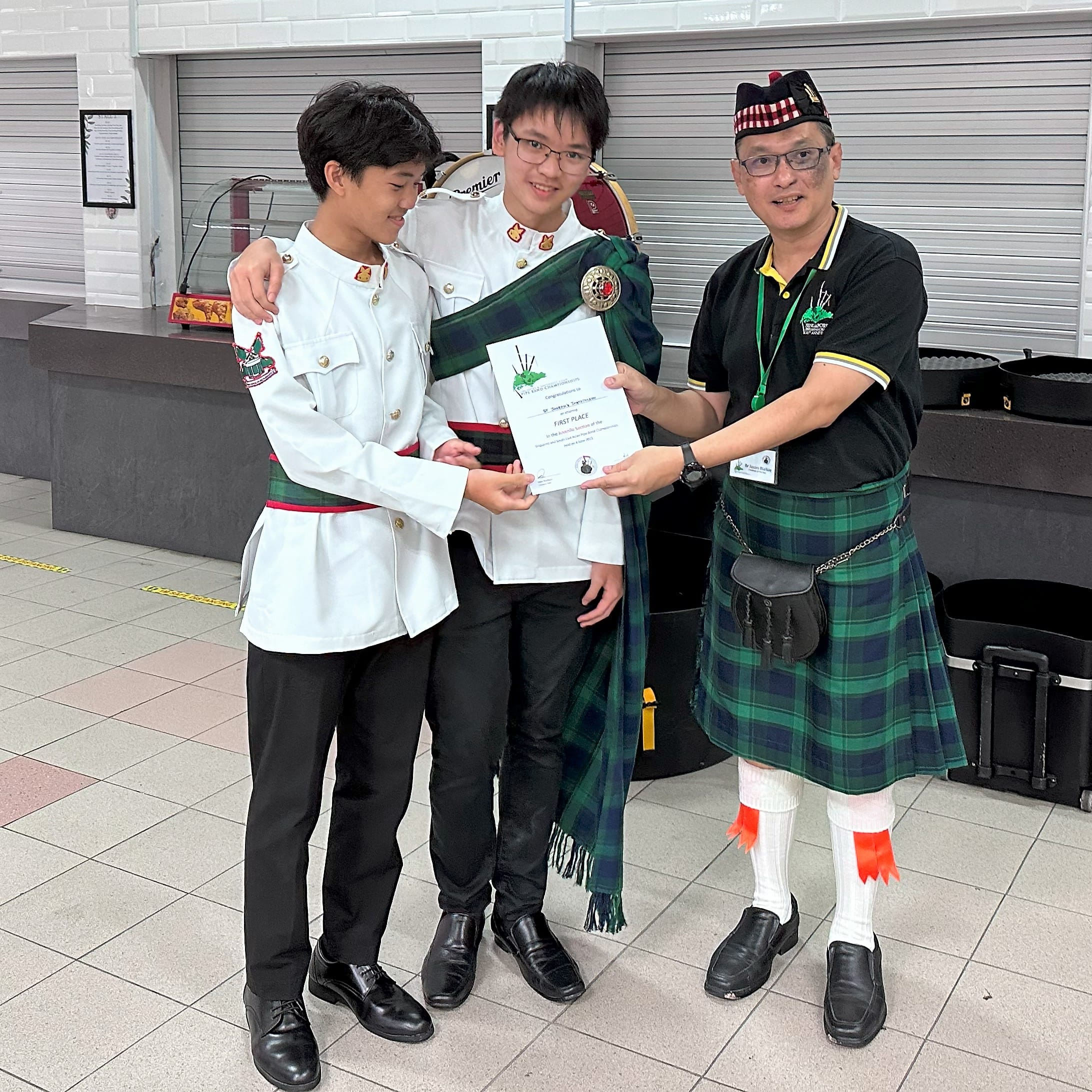 Three people pose indoors holding a First Place certificate; two wear white uniforms and one wears a green tartan kilt.