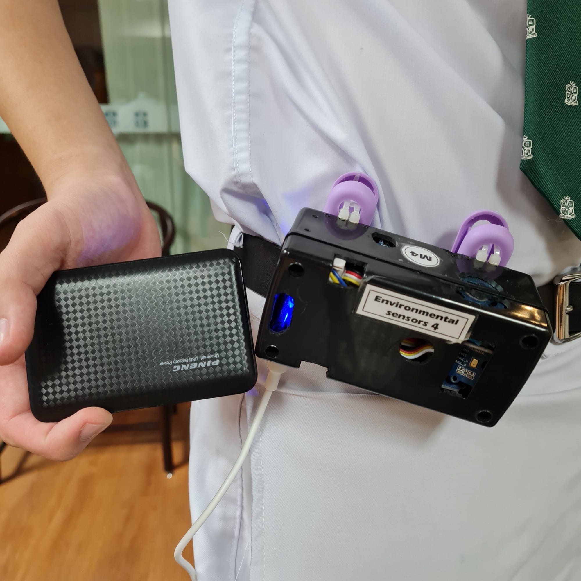 A hand holds a Pineng power bank connected via USB to a black box labeled "Environmental sensors 4" clipped to a belt.