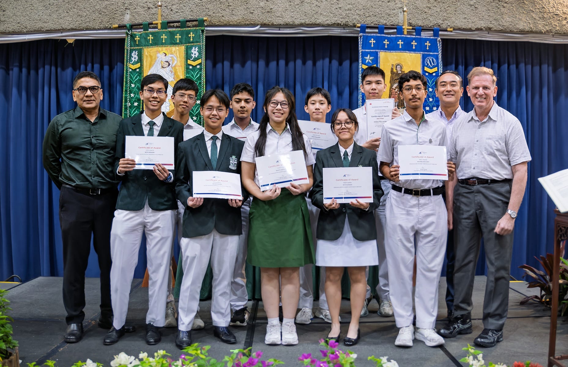 A group of students and staff pose on stage holding certificates of award in front of decorative banners at a school ceremony.