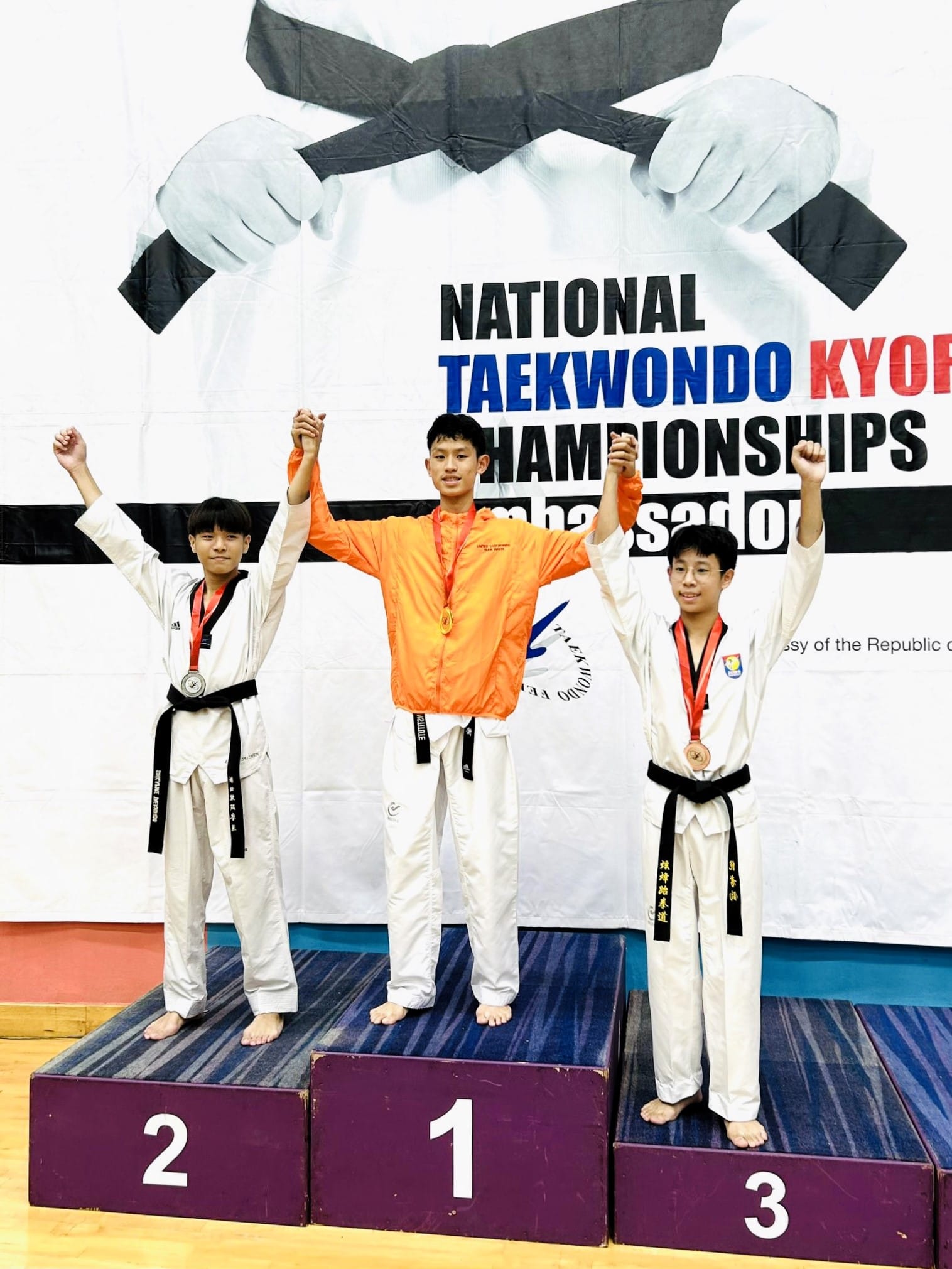 Three young taekwondo competitors stand on a podium with raised arms at the National Taekwondo Kyorugi Championships.