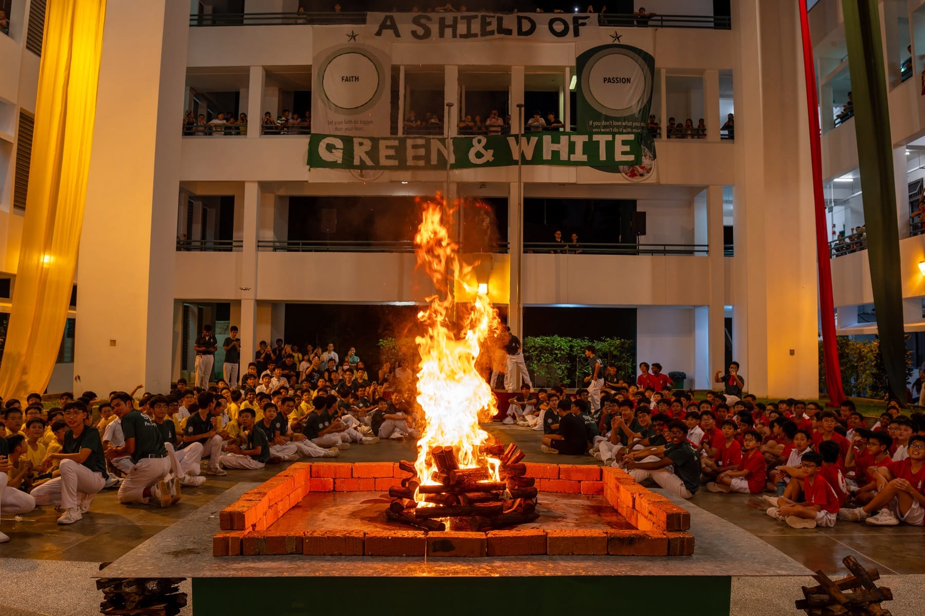 Students in colored uniforms sit around a large bonfire at night, with a banner reading "A Shield of Green & White."