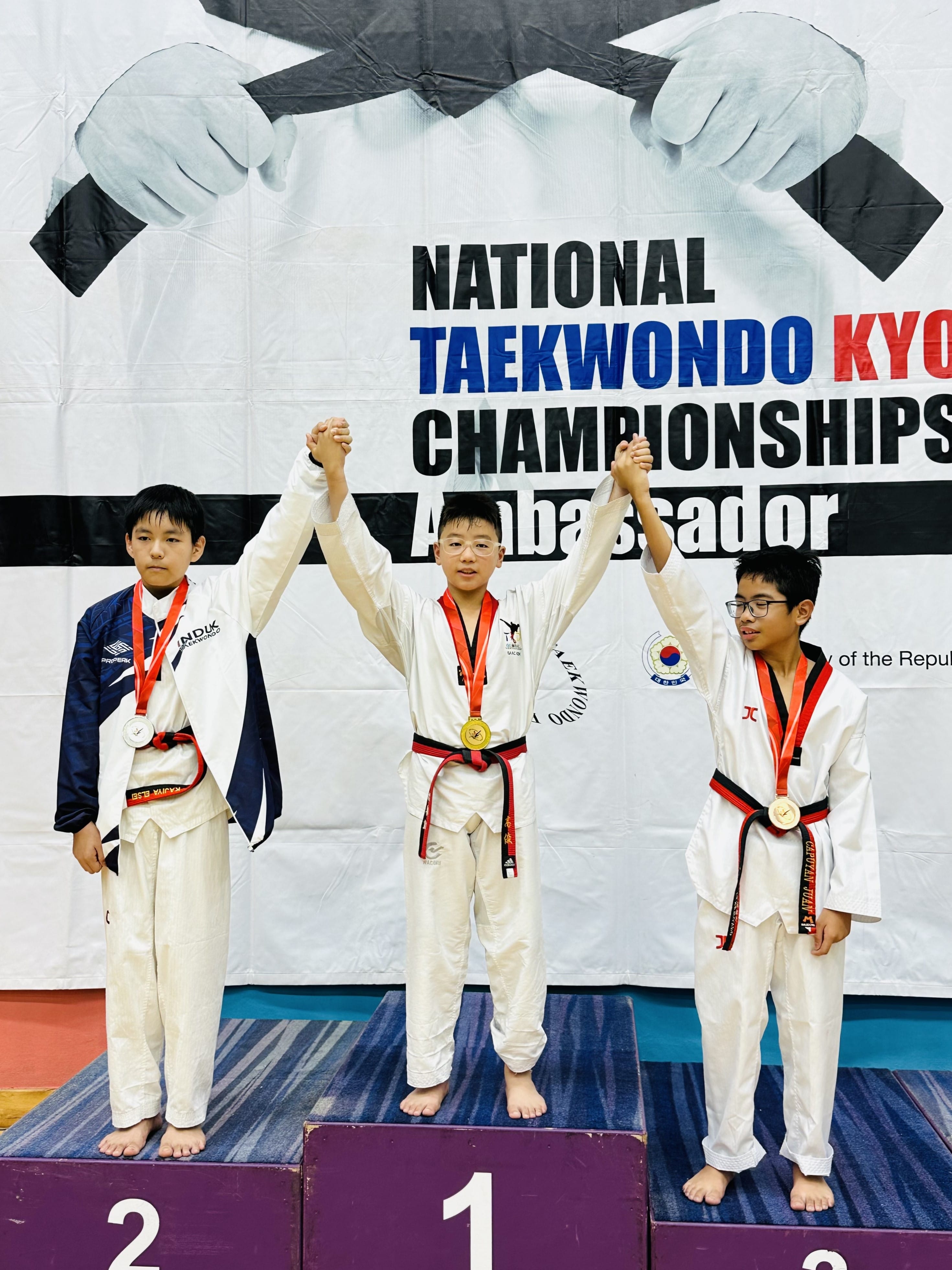 Three young taekwondo boys in dobok uniforms stand on a podium with medals at a National Taekwondo Championship.