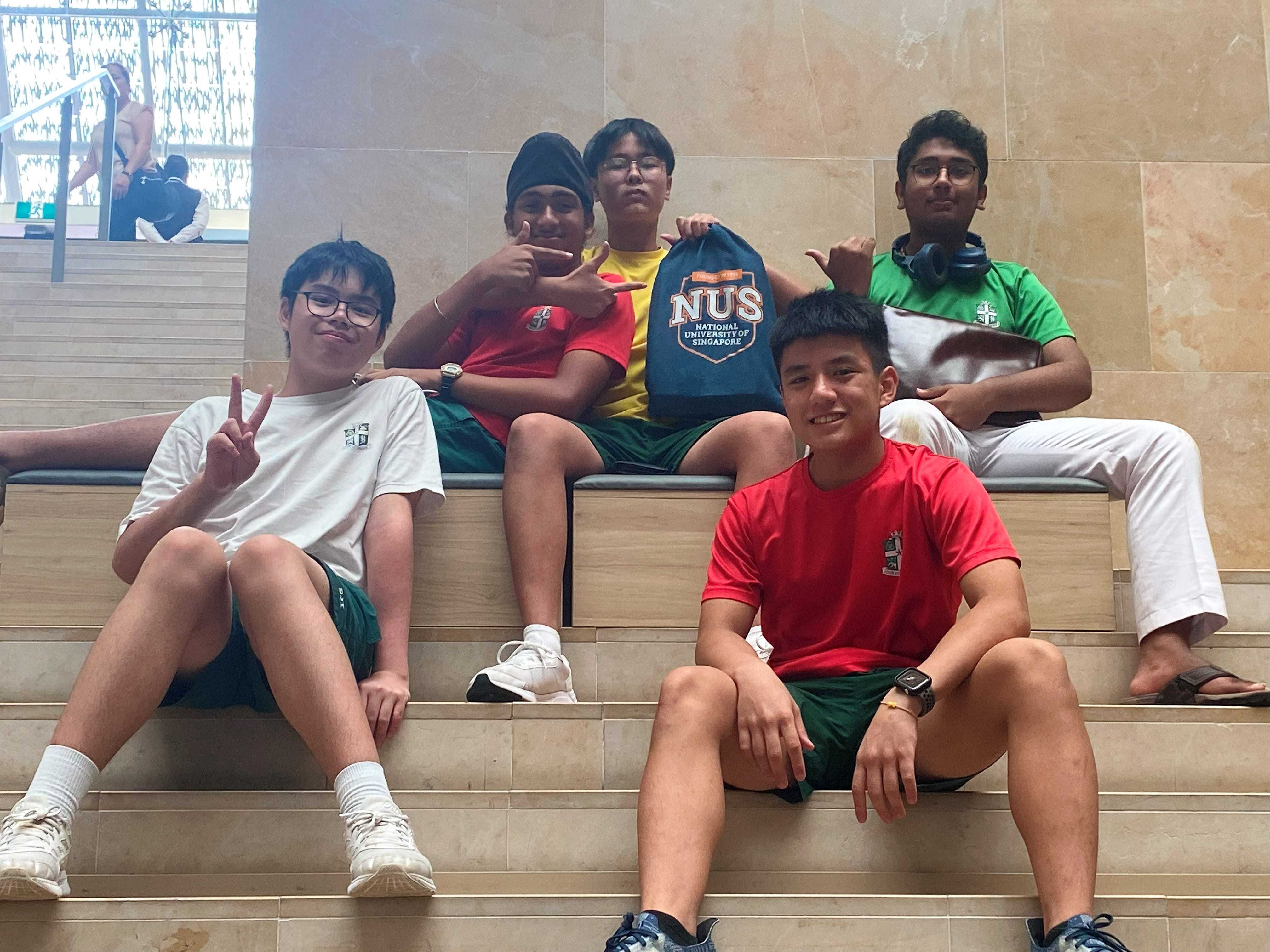 Students posing on steps inside a marble-walled hall, one holding an NUS drawstring bag.