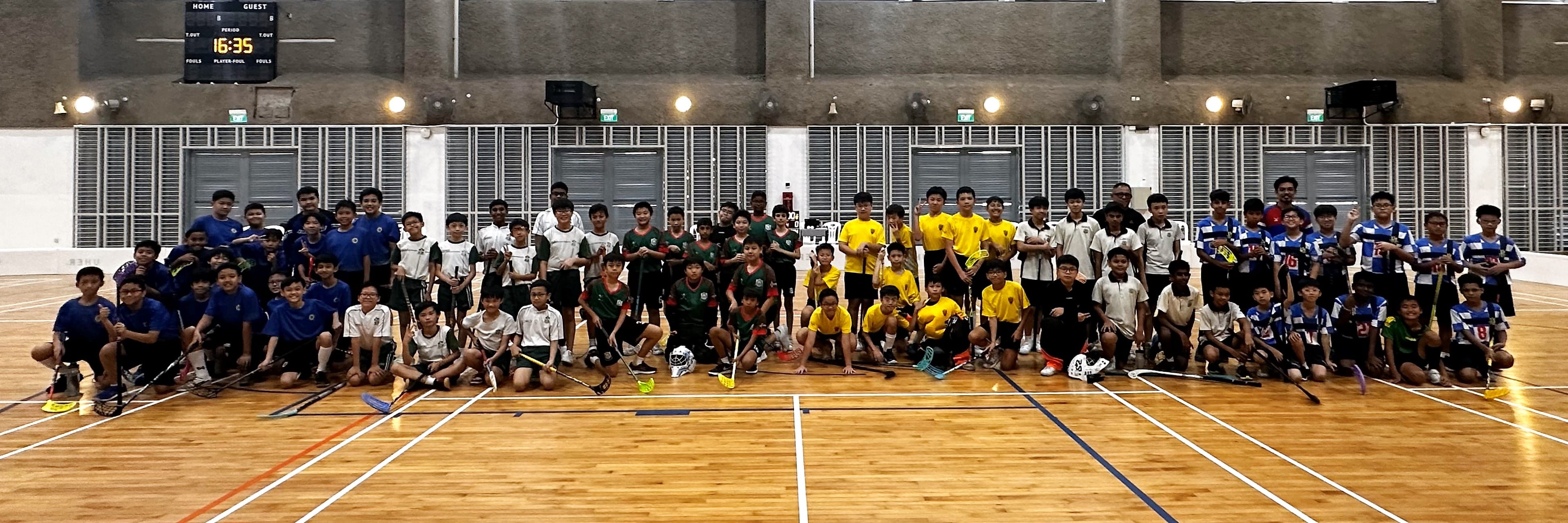 Large group of youth floorball players in colorful jerseys posing with sticks on an indoor wooden court.