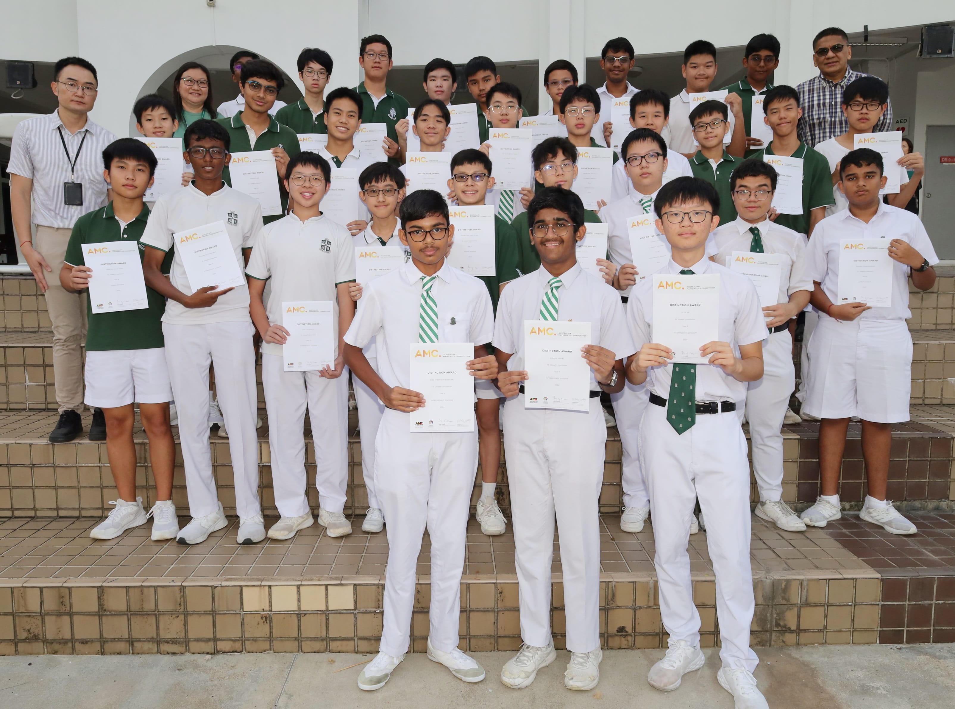 A large group of school boys in white and green uniforms holding AMC distinction award certificates on outdoor steps.