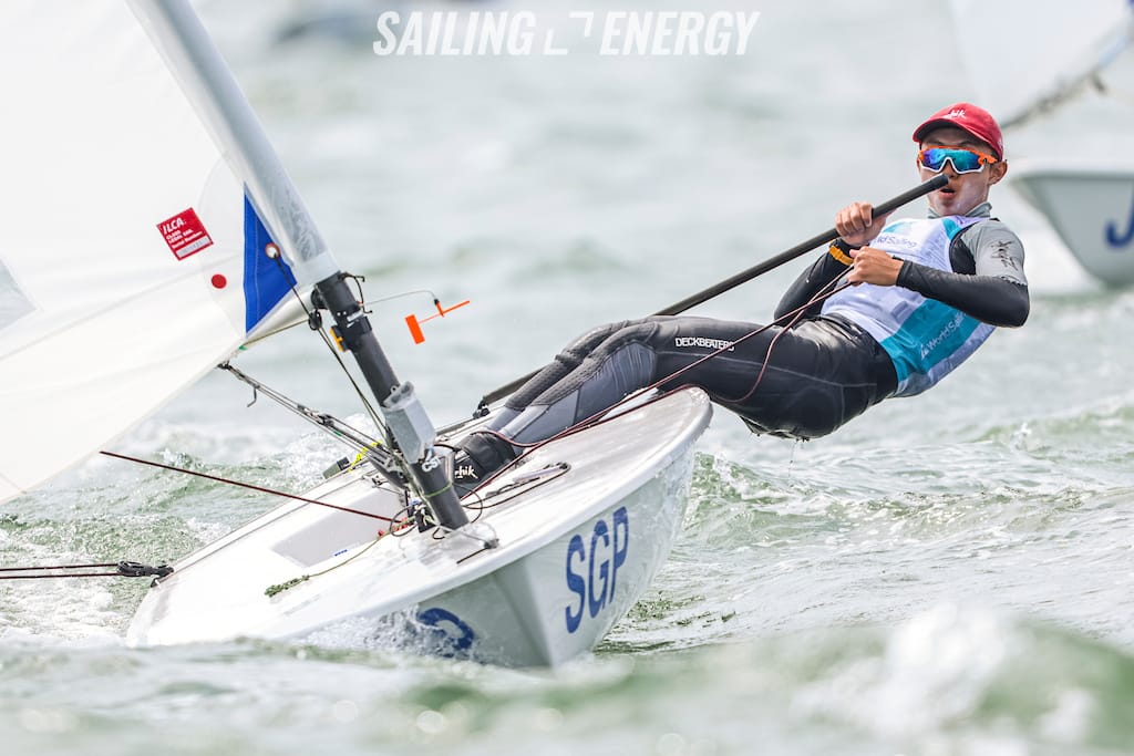 A sailor in a wetsuit and red cap leans back on a small SGP-marked dinghy racing on choppy water.
