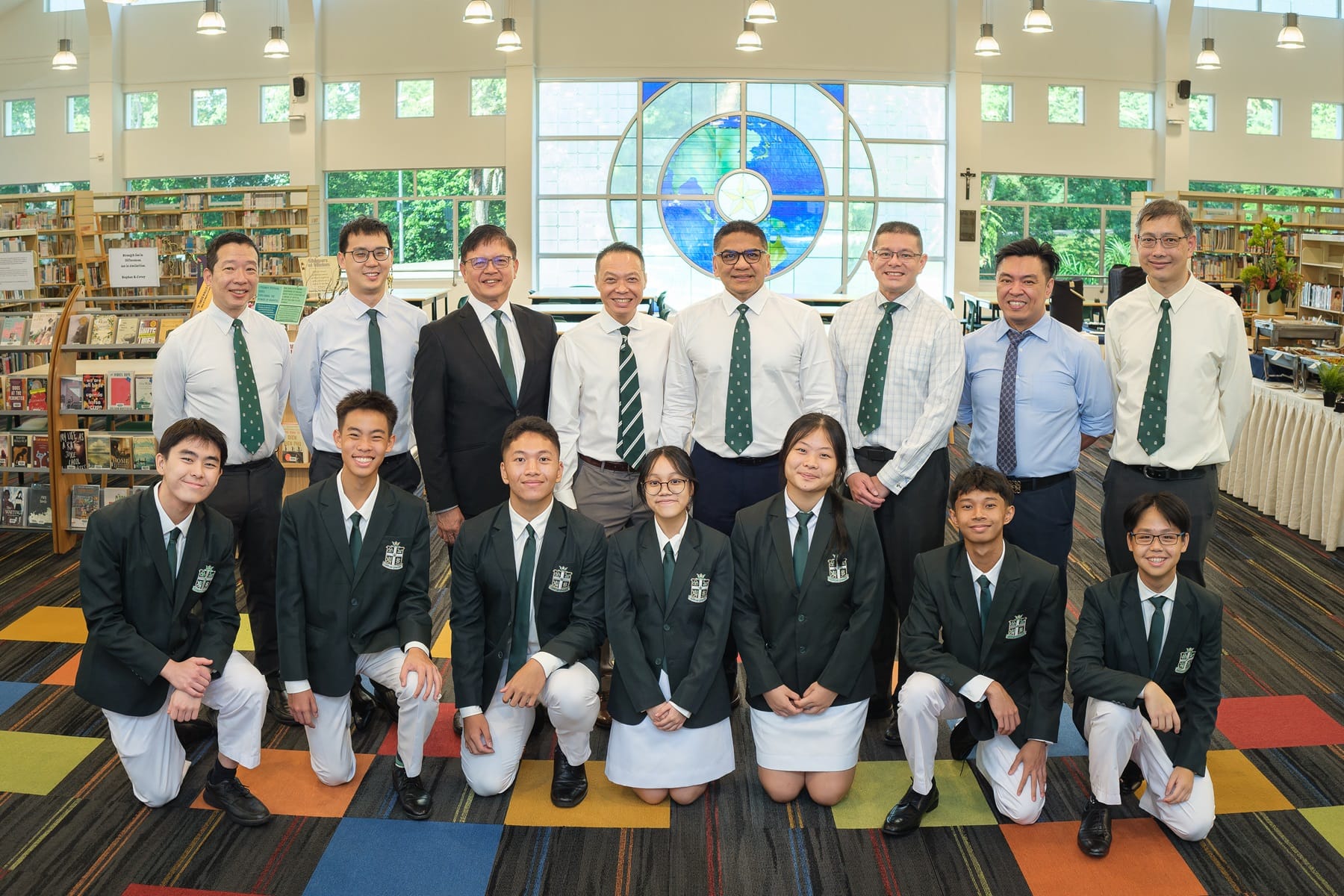 Group of students in dark blazers and teachers in white shirts pose in a school library with a stained glass window.