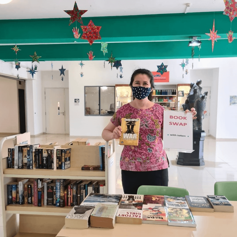 Woman wearing a mask holds a book and a "Book Swap" sign beside shelves of books, with star decorations above.