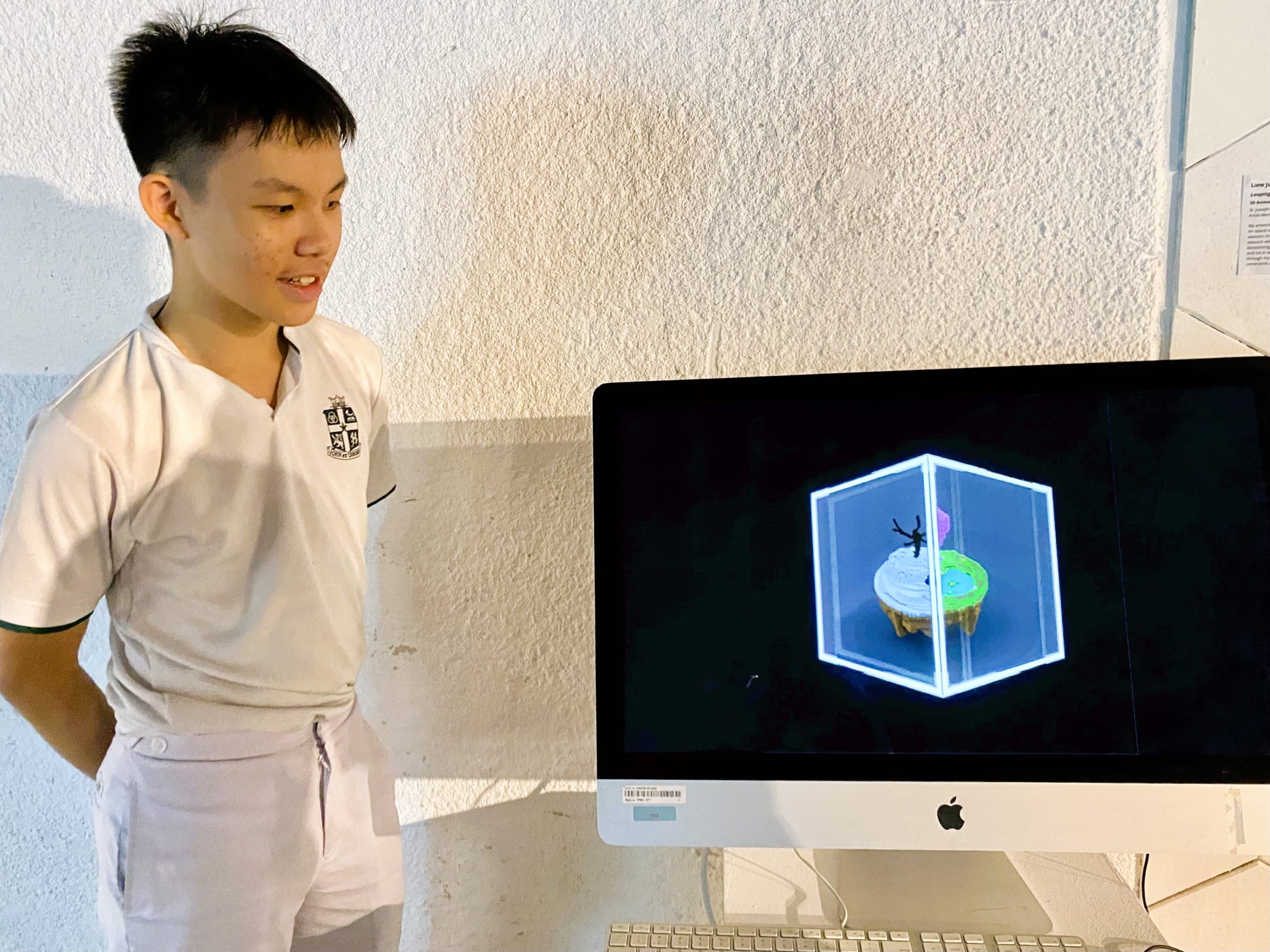 A young male student in white uniform stands beside an iMac displaying a 3D animated cupcake inside a glowing cube.
