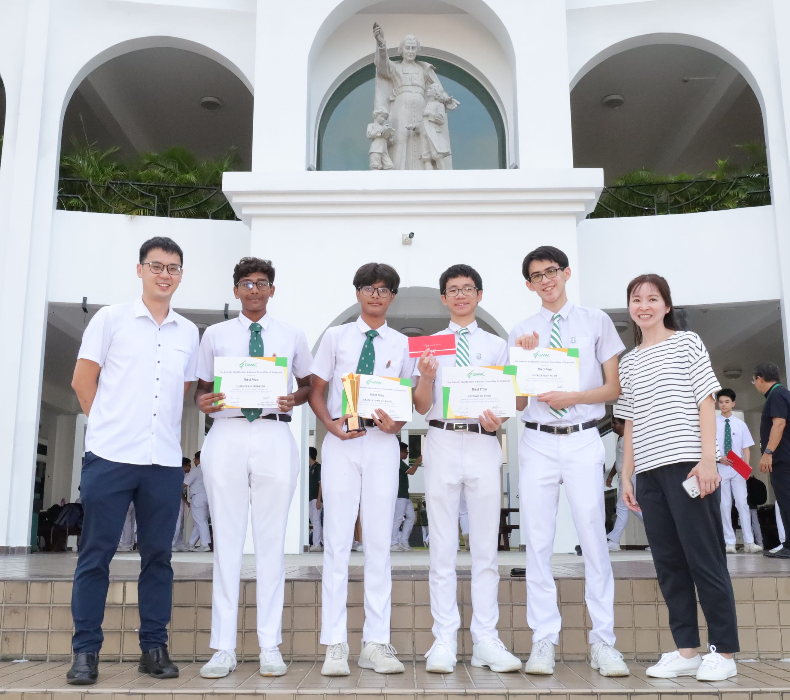 Six people pose outdoors holding certificates and a trophy in front of a white building with a statue.