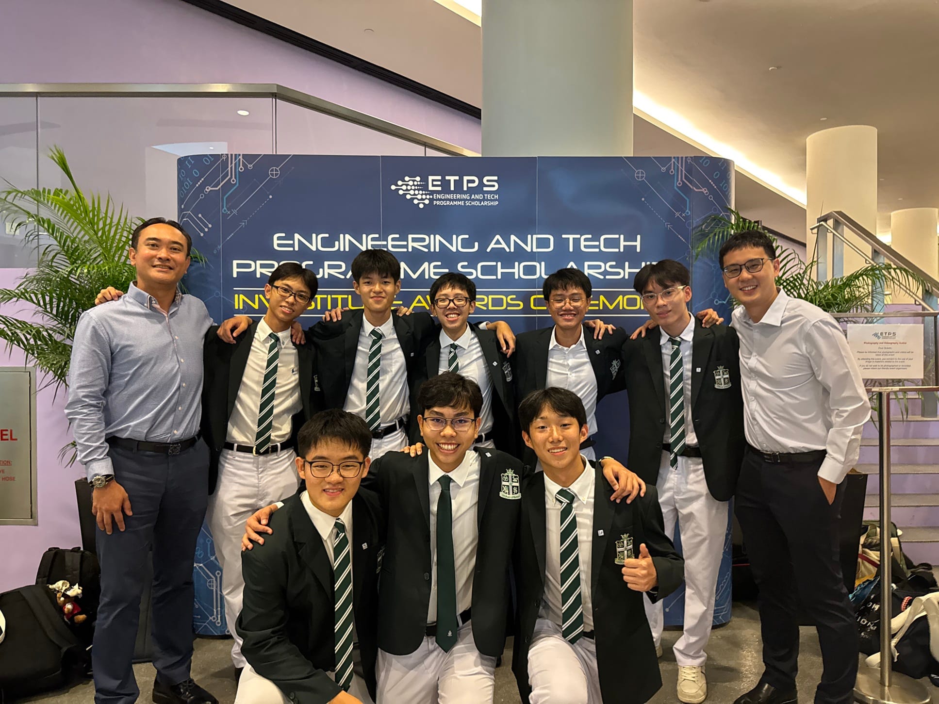 Group of students in school uniforms posing with two adults before an ETPS Engineering and Tech Programme Scholarship banner.