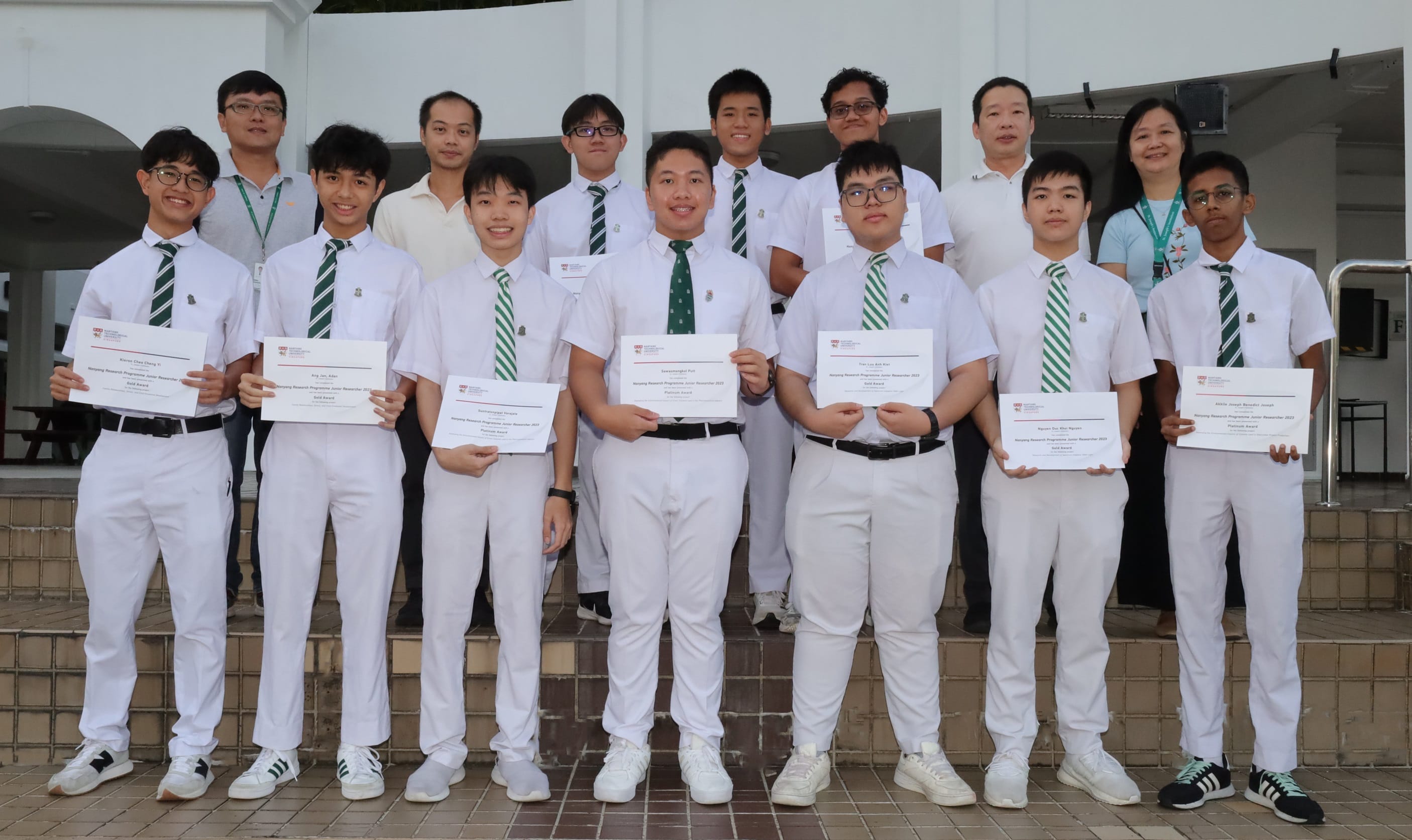 Students in white uniforms with green striped ties hold award certificates while posing in two rows outdoors.