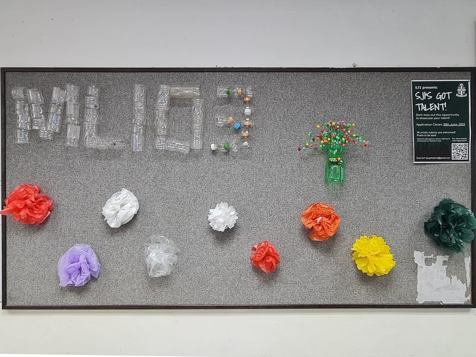 Bulletin board spelling MILO3 in plastic bottles, with tissue-paper flowers below and a poster for SJI's Got Talent at right.