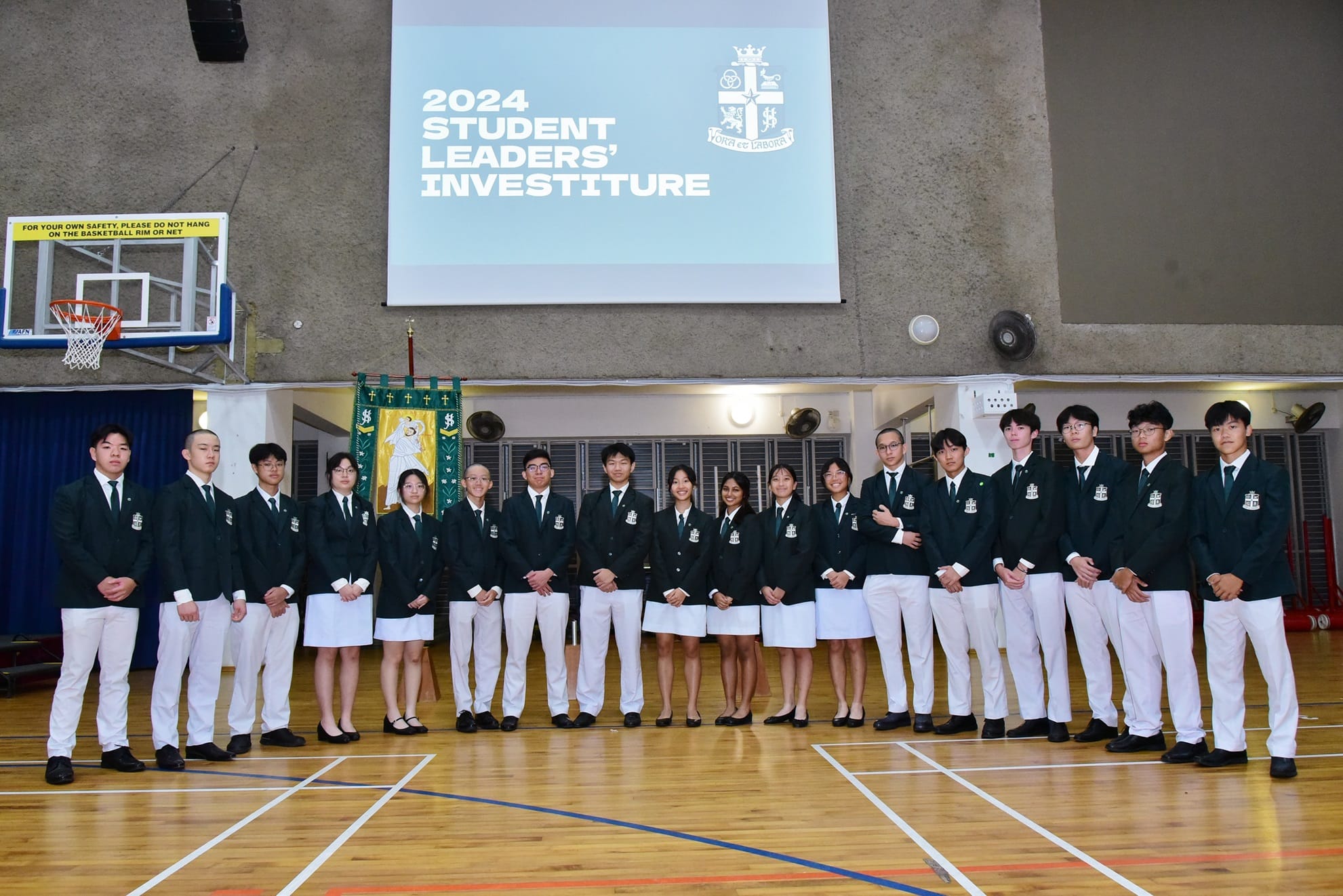 Students in dark blazers and white bottoms pose in a gym under a "2024 Student Leaders' Investiture" projection screen.