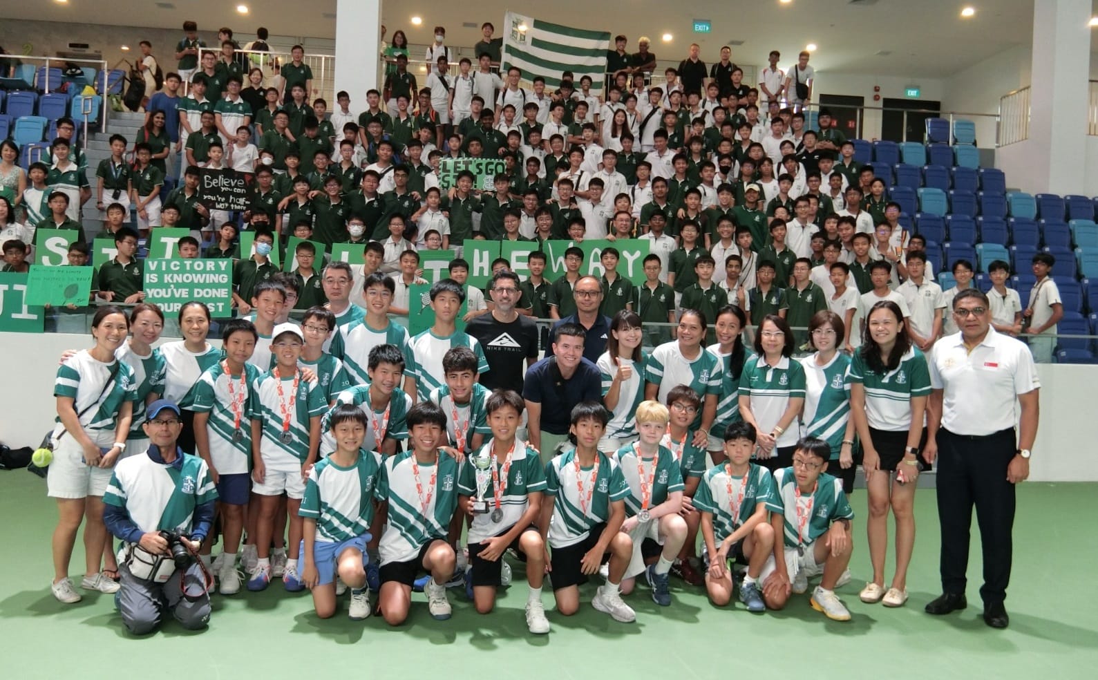 Large group of students in green and white uniforms posing with a trophy, cheering crowd holding signs in bleachers behind.