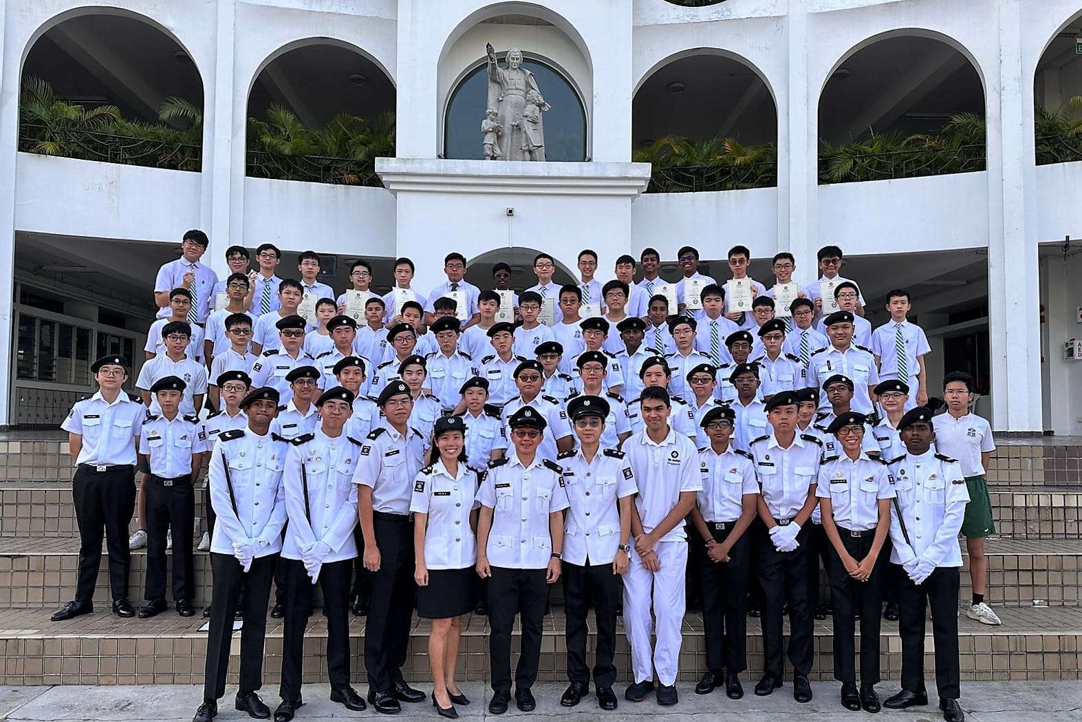 Large group of students in white uniforms and berets posing on steps in front of a white building with a statue.