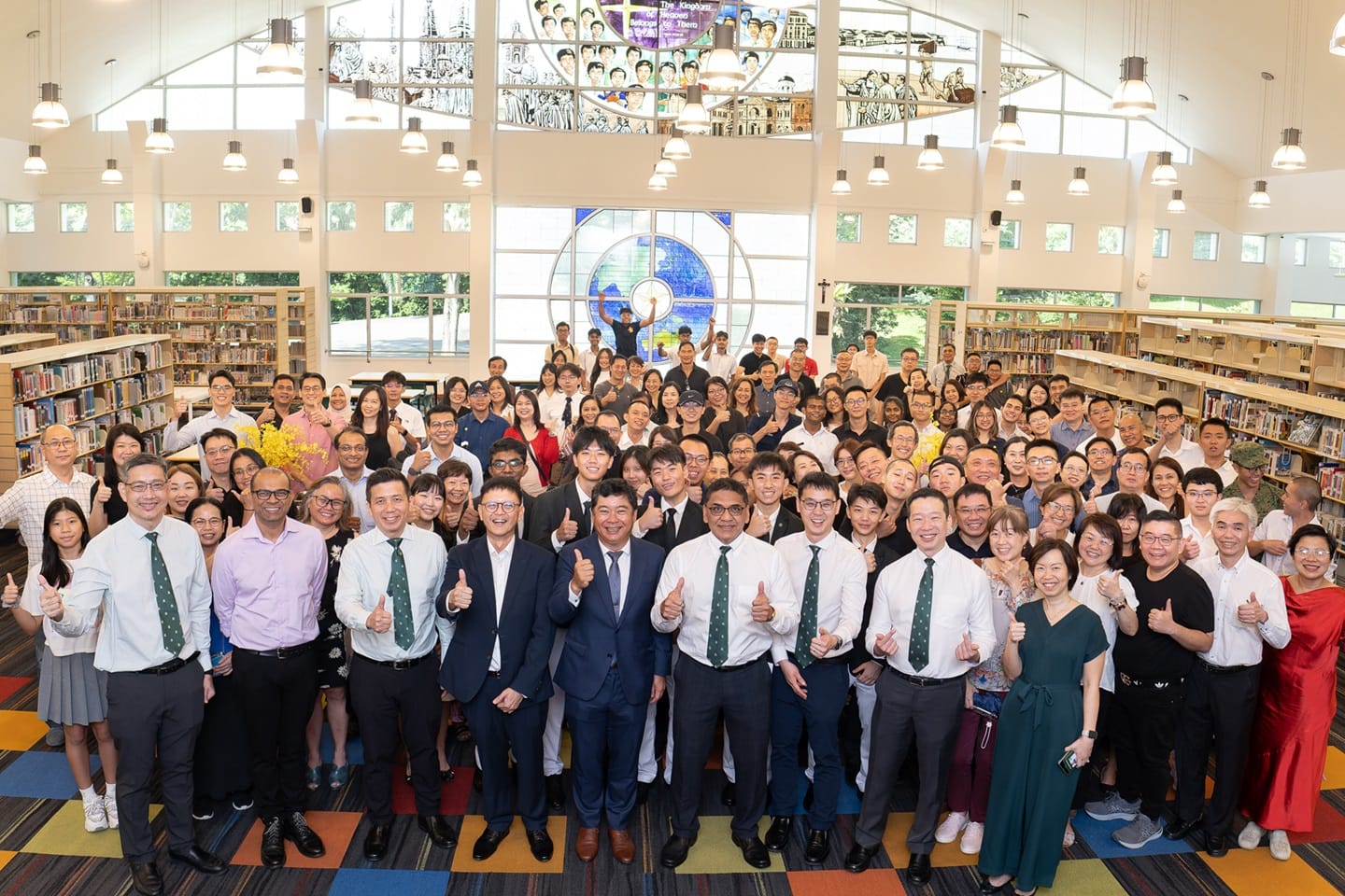 Large group of people posing with thumbs up in a bright library with bookshelves and a stained glass window.