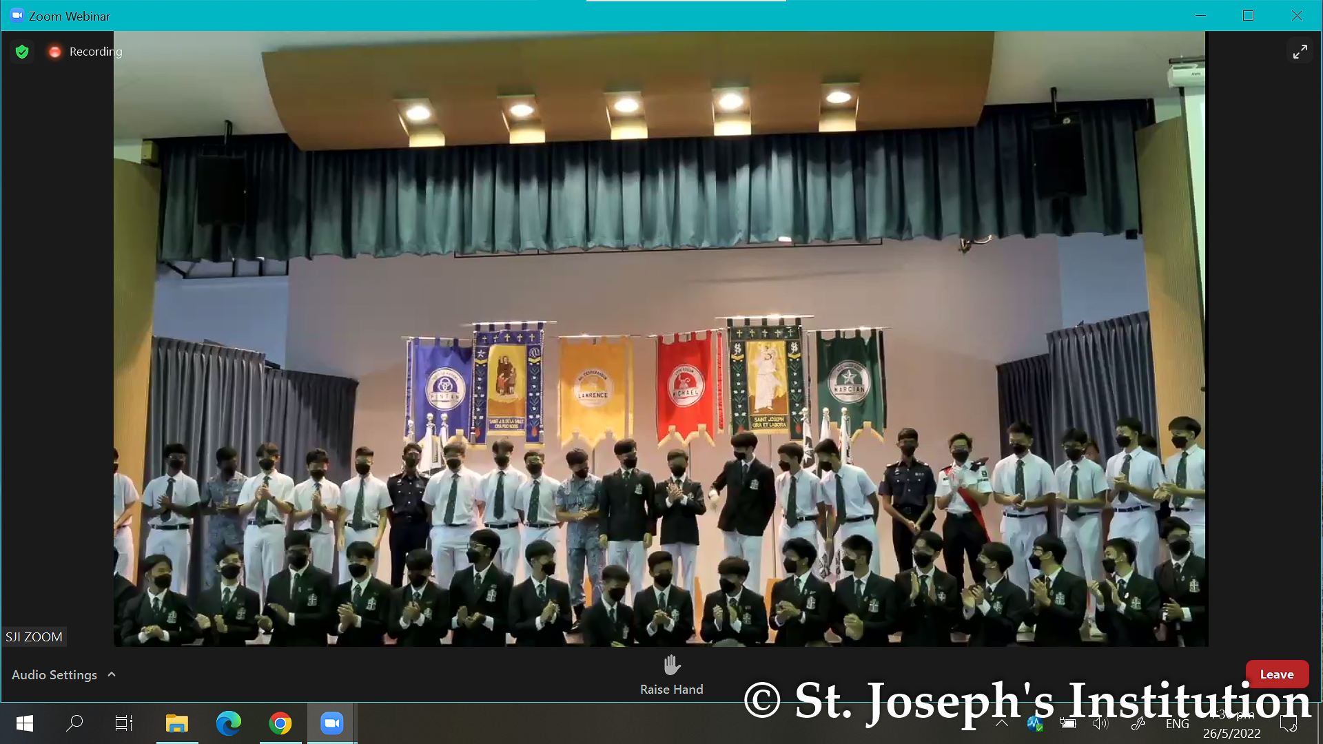 Masked male students in uniforms stand on a stage with colorful banners during a Zoom webinar at St. Joseph's Institution.