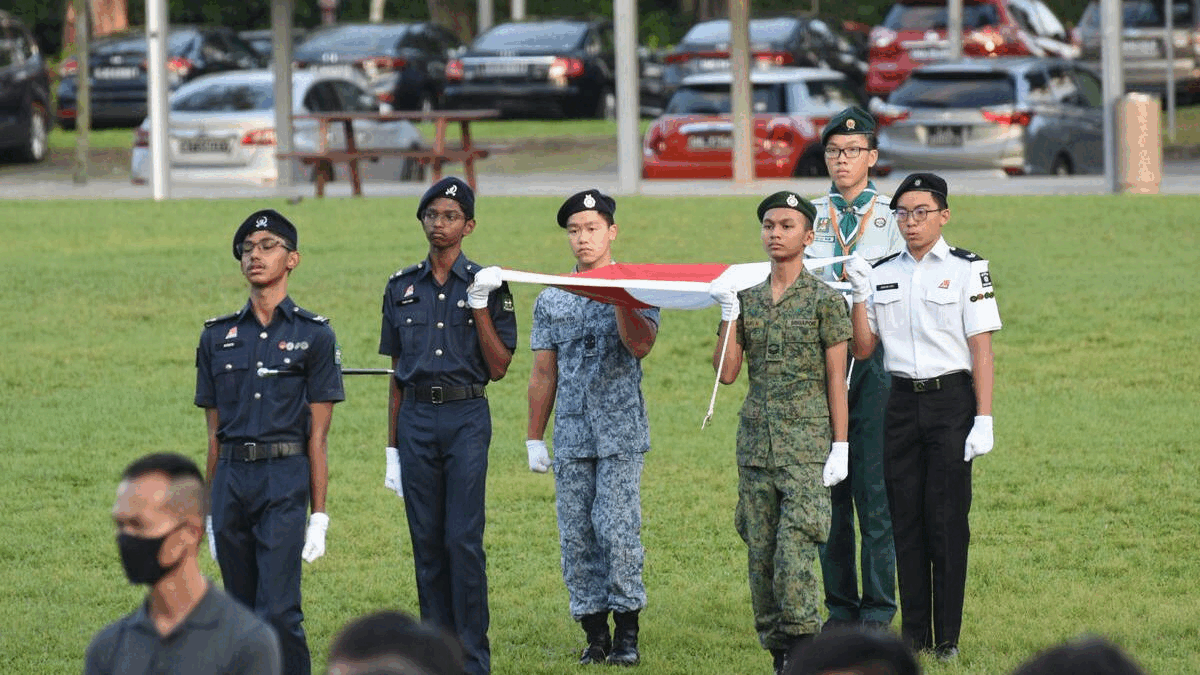 Six young people in various uniforms hold a red and white flag on a grass field during a ceremony.