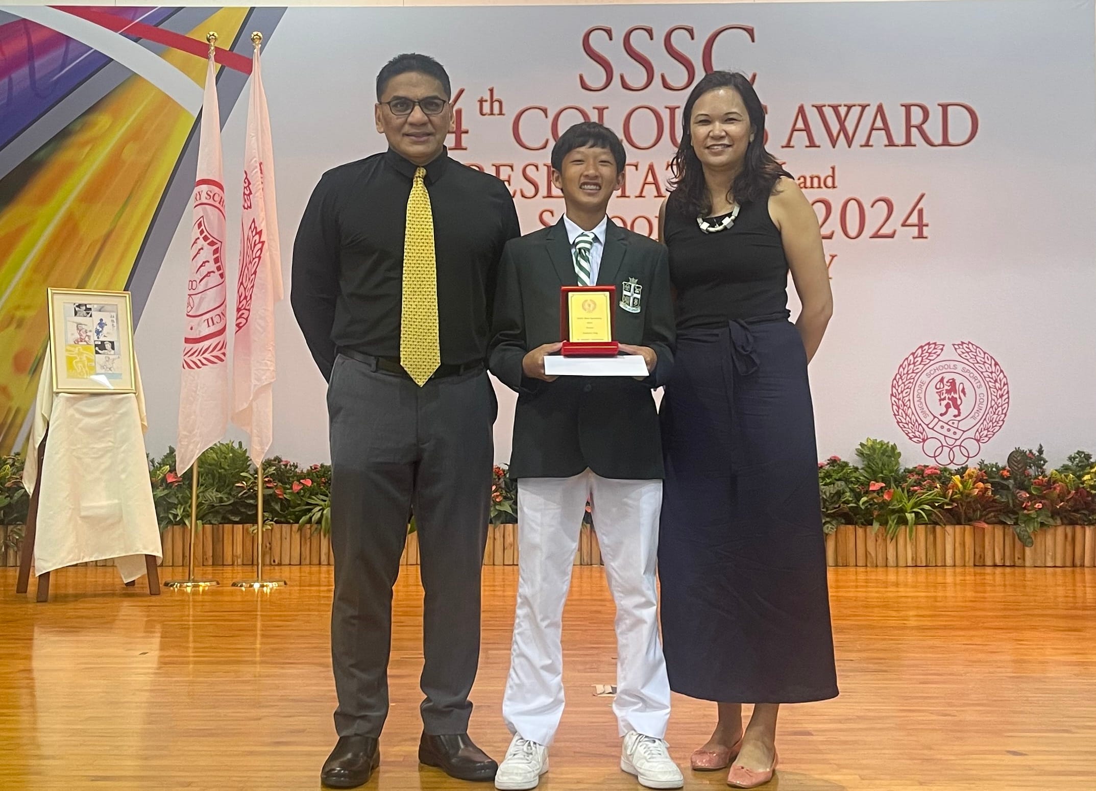 A smiling student in a green blazer holds a plaque flanked by two adults at the SSSC Colours Award 2024.