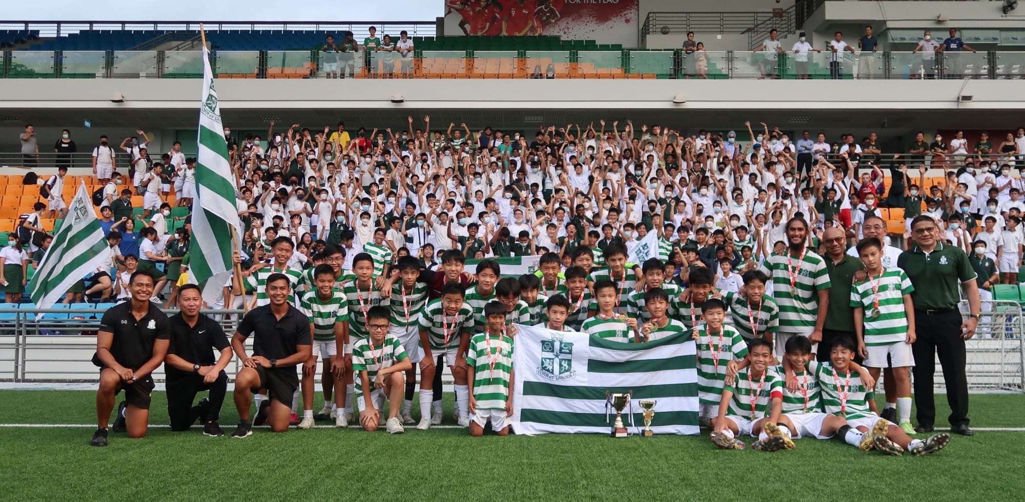 Youth football team in green and white striped kits poses with trophies and a flag before a cheering crowd in a stadium.