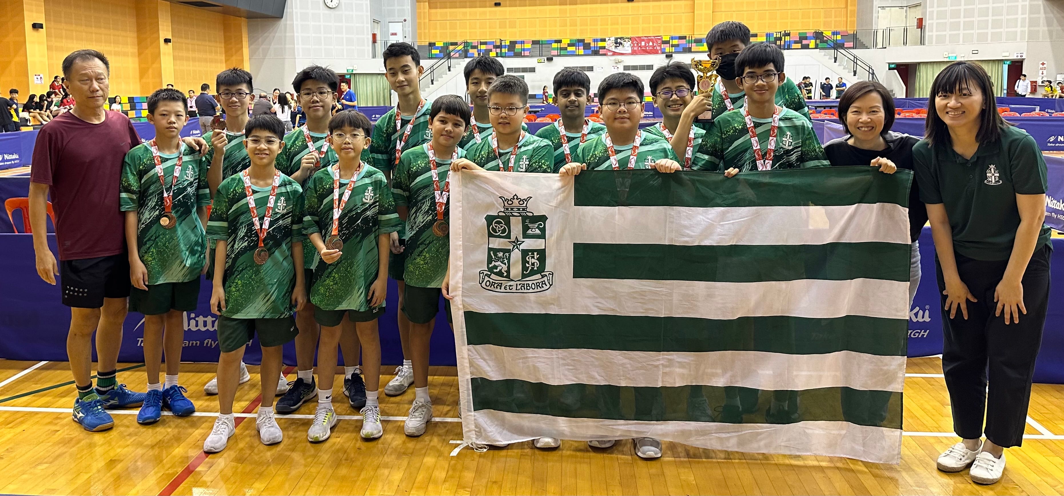 A group of boys in green uniforms with medals hold a green and white striped school flag and a trophy in a sports hall.