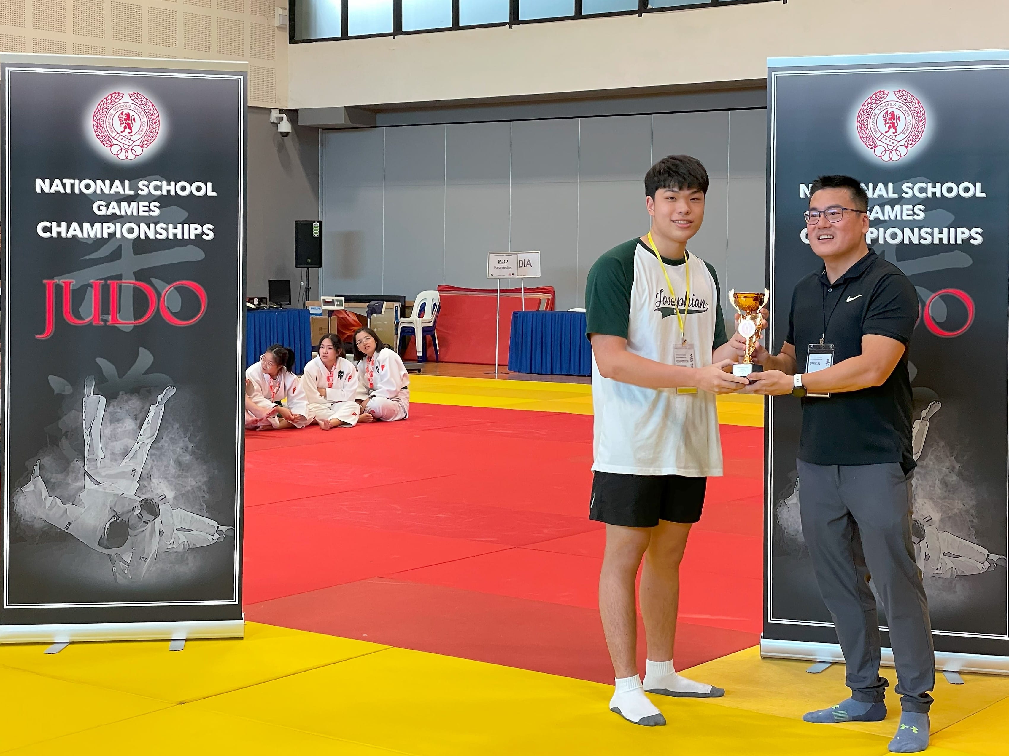 A student athlete in sportswear receives a trophy from an official beside National School Games Championships Judo banners, with judokas seated in the background.