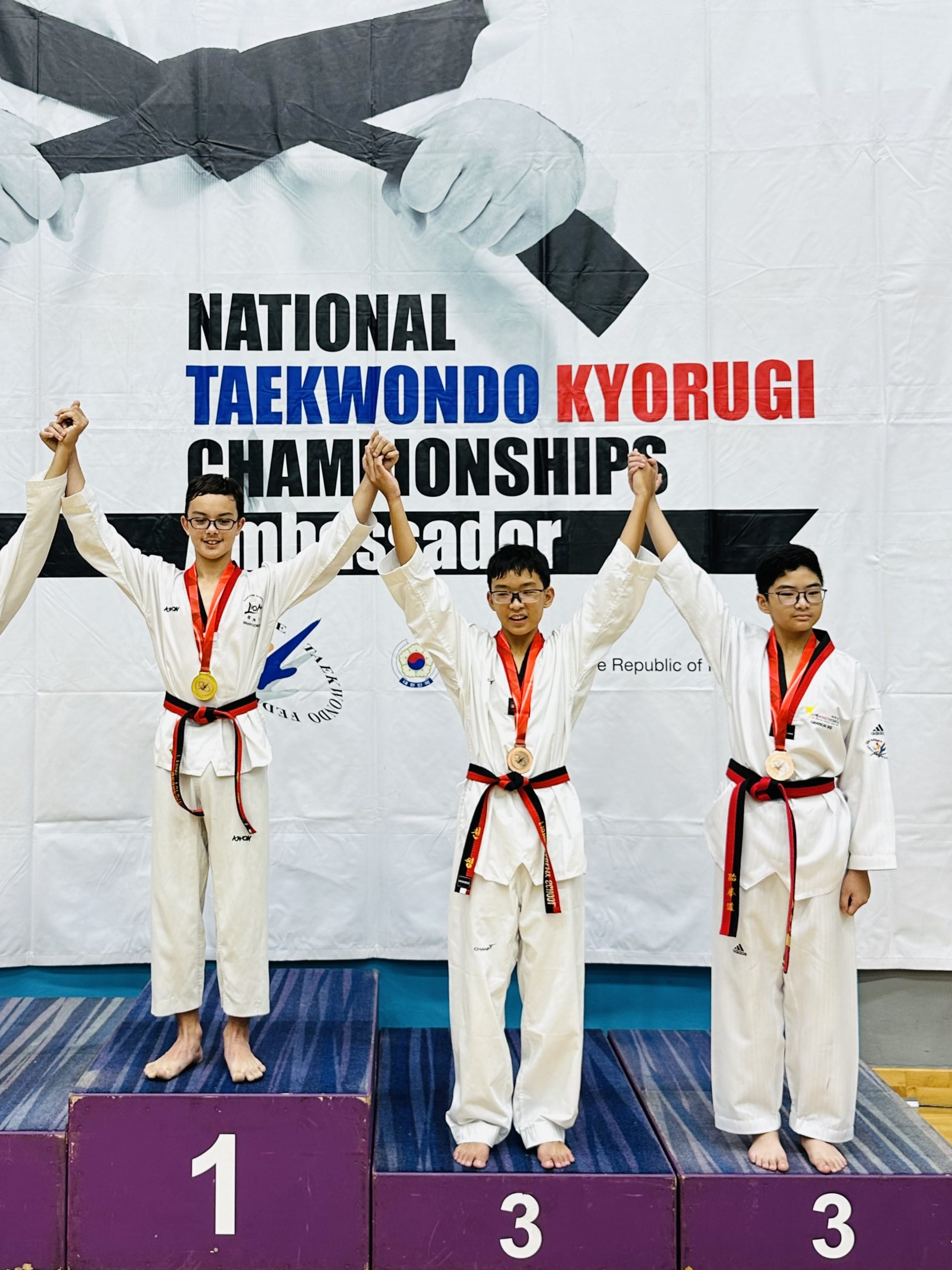Three young taekwondo competitors in white uniforms with medals stand on a podium at the National Taekwondo Kyorugi Championships.