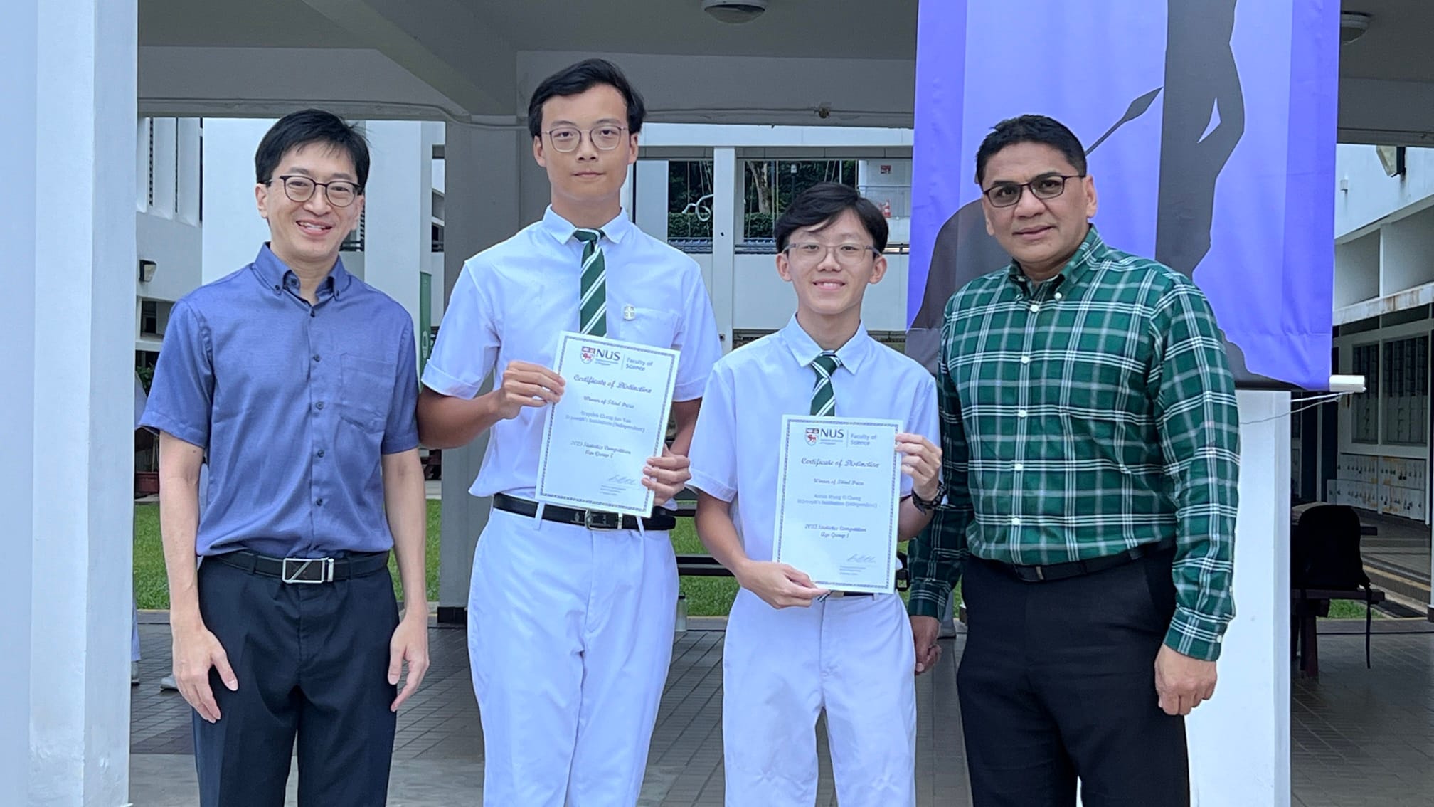 Four people stand outdoors; two students in white uniforms hold NUS Certificates of Distinction while two adults stand beside them.