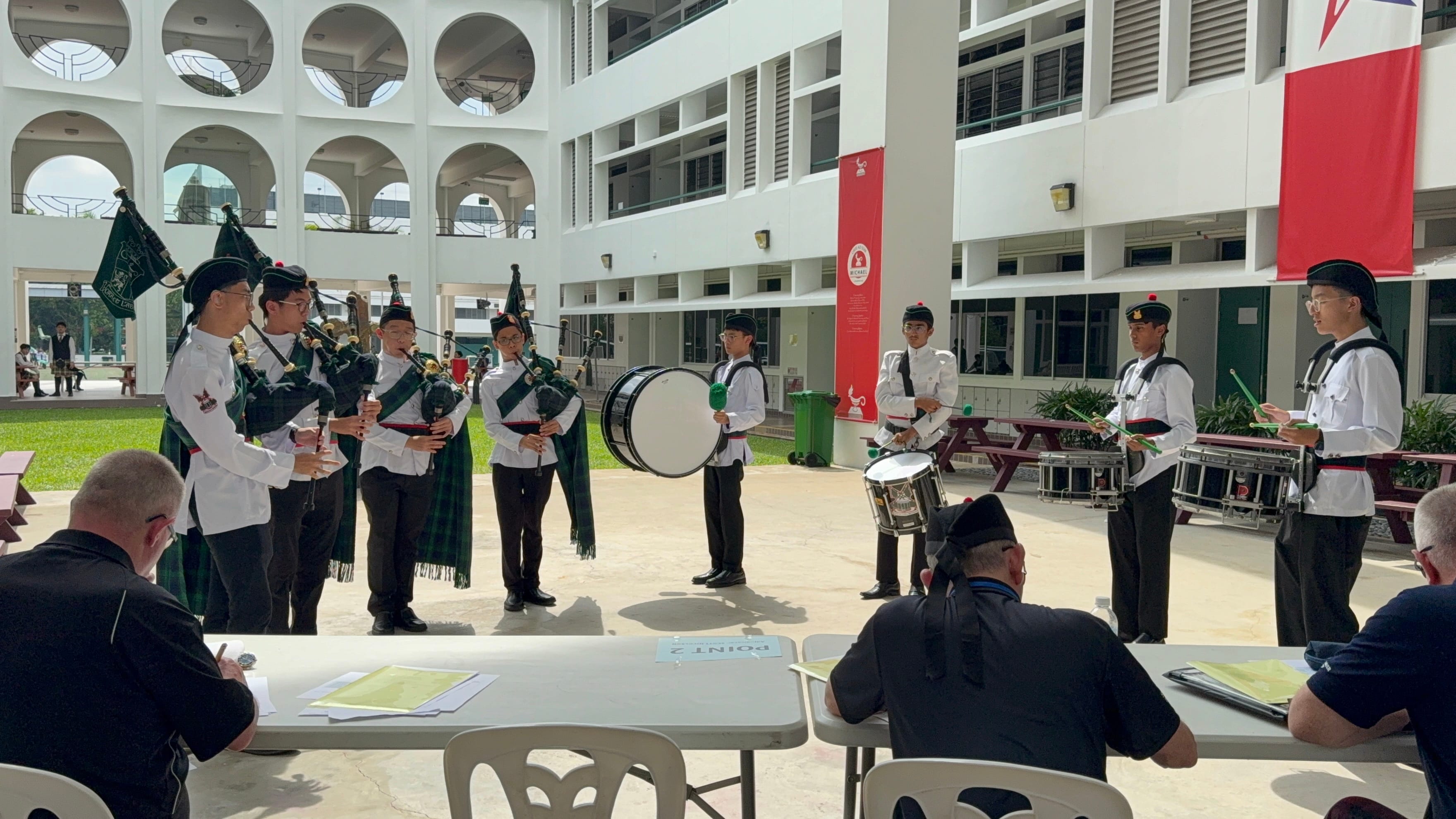 Students in Highland uniforms playing bagpipes and drums perform for judges seated at a table in a courtyard.