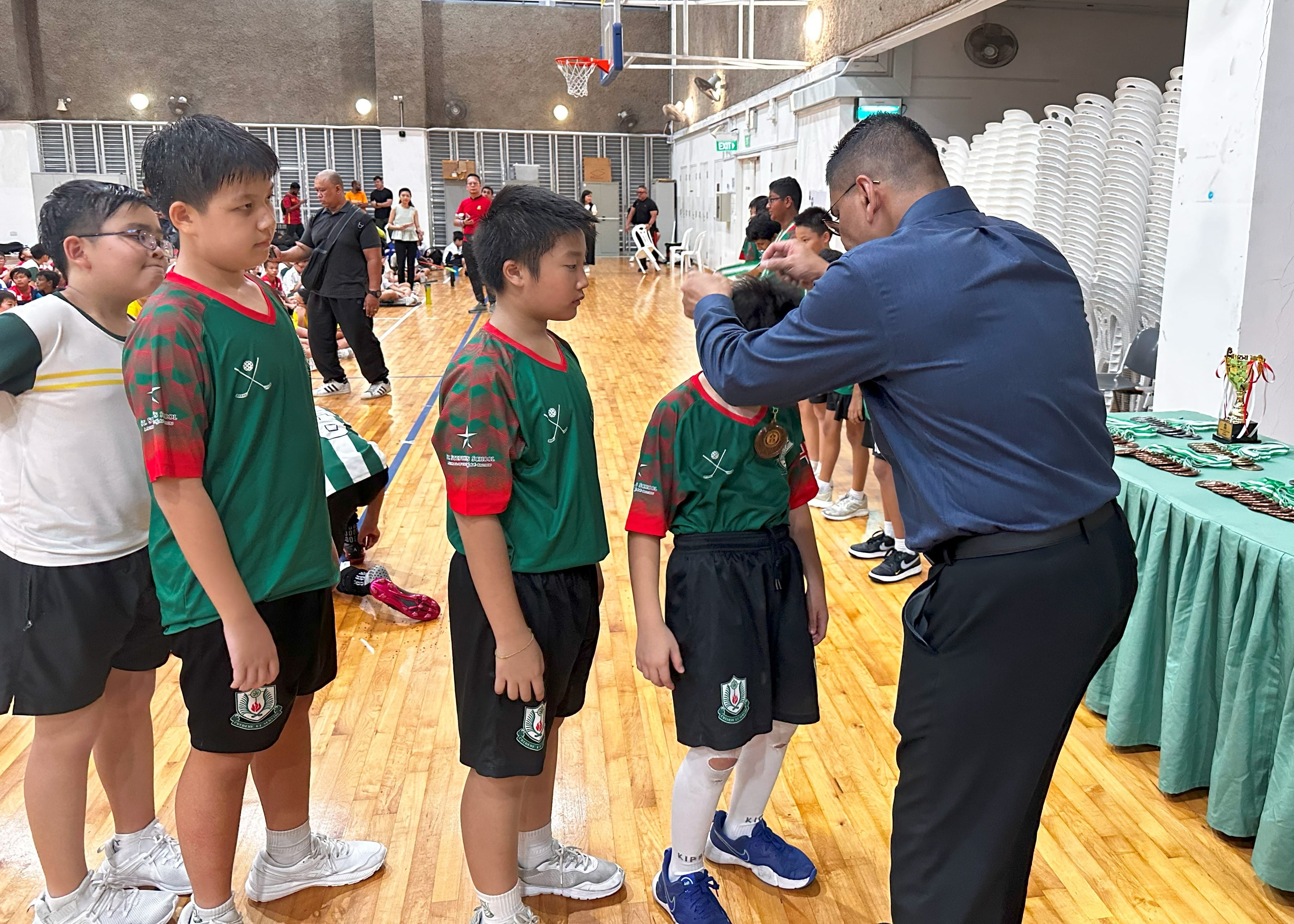 A man in a blue shirt places a medal around a boy's neck as other boys in green jerseys wait in line indoors.