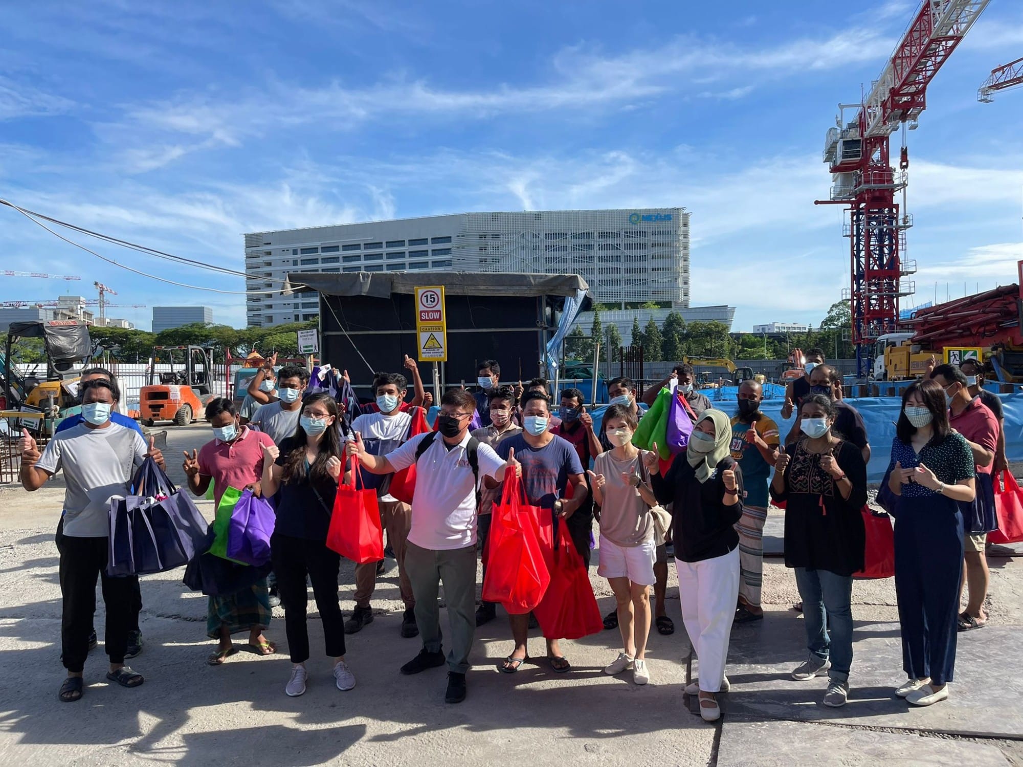 A group of masked people holding colorful tote bags give thumbs up at an outdoor construction site with a crane and building behind them.