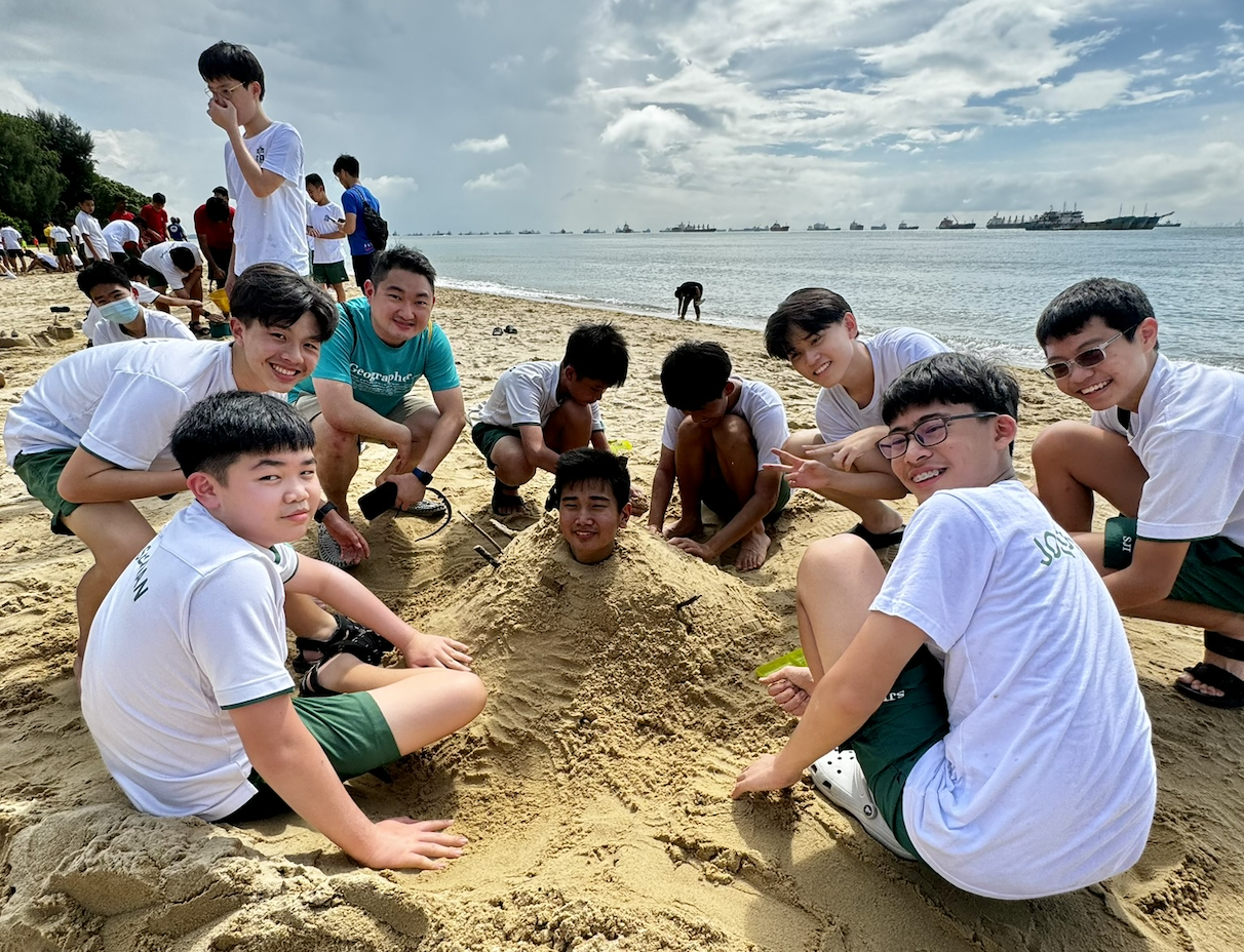 A group of smiling teenagers on a beach surround a person buried in sand with only their head visible.