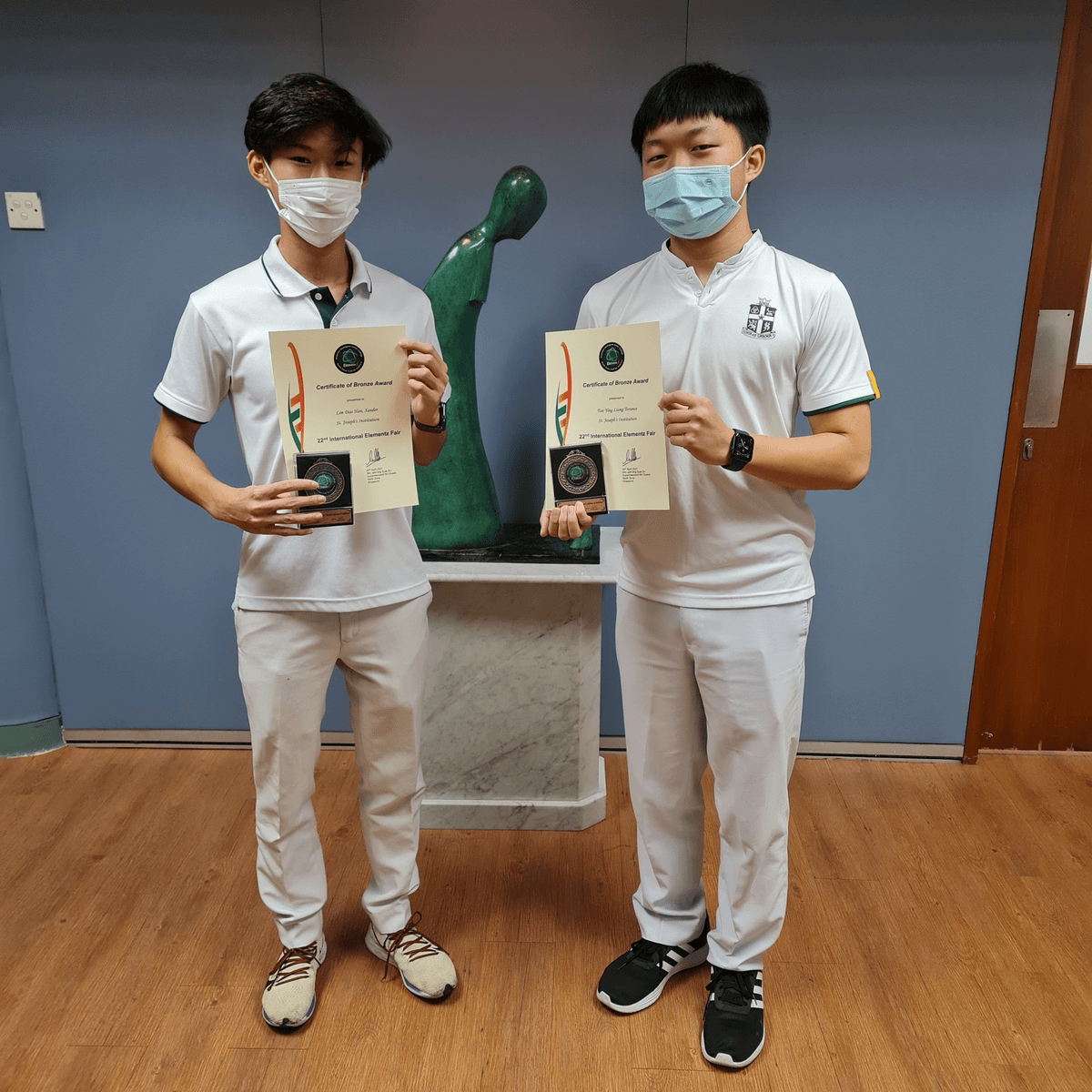 Two masked students in white uniforms hold Certificate of Bronze Award plaques from the 22nd International Elements Fair.