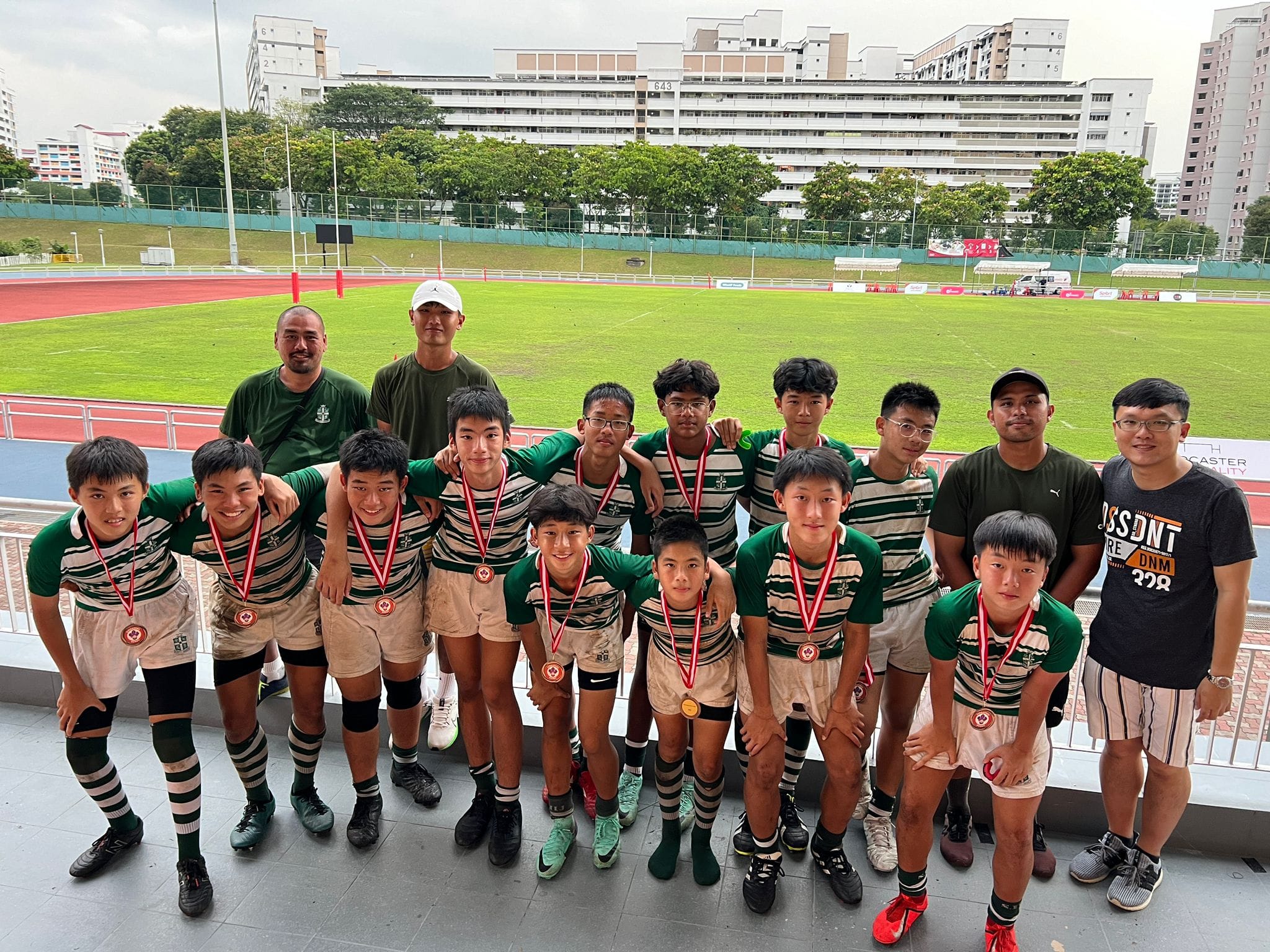A youth rugby team in green striped uniforms poses with medals and coaches at a sports stadium.