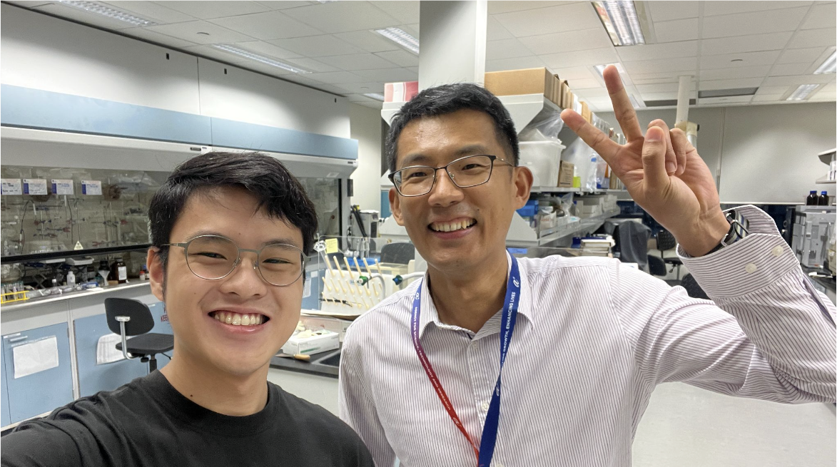 Two smiling men wearing glasses pose for a selfie in a science laboratory, one making a peace sign.