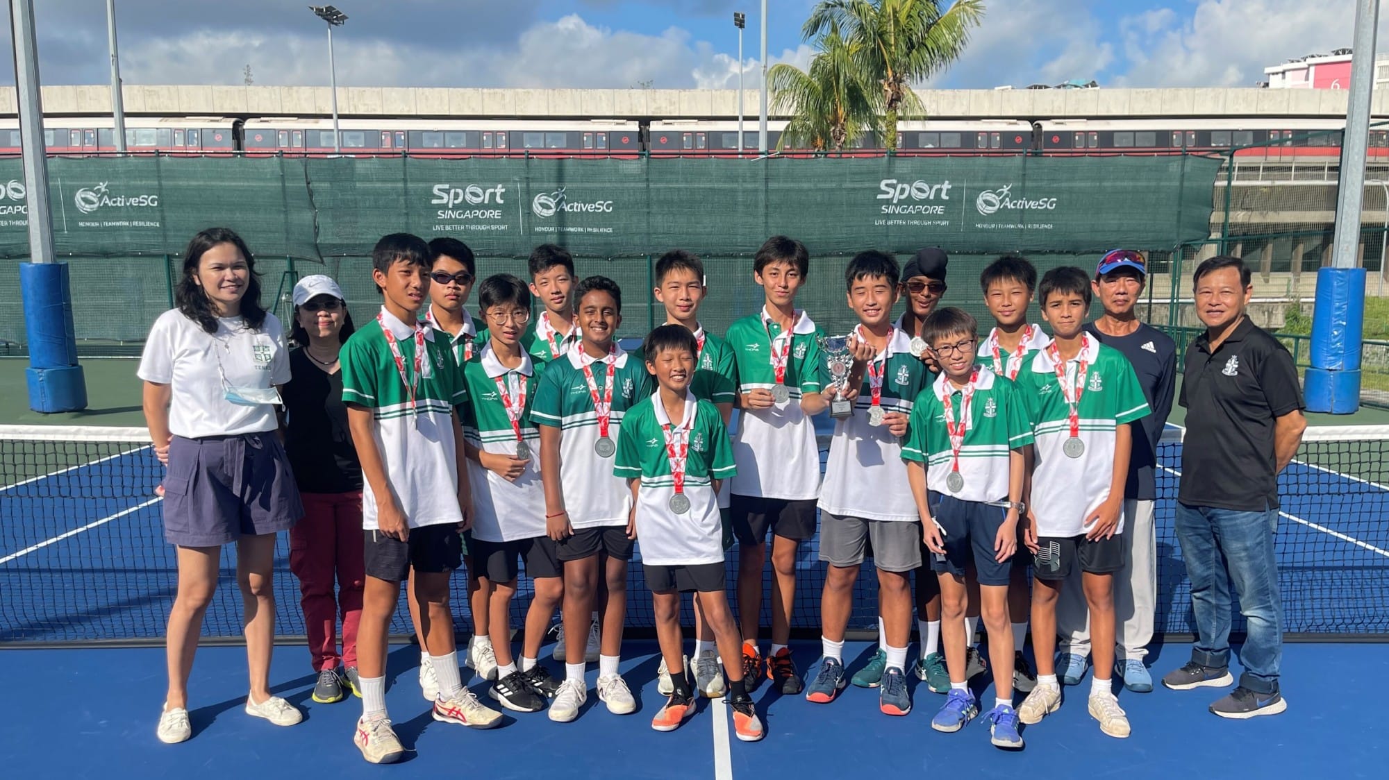A youth tennis team in green uniforms poses with medals and trophies on a Sport Singapore court.