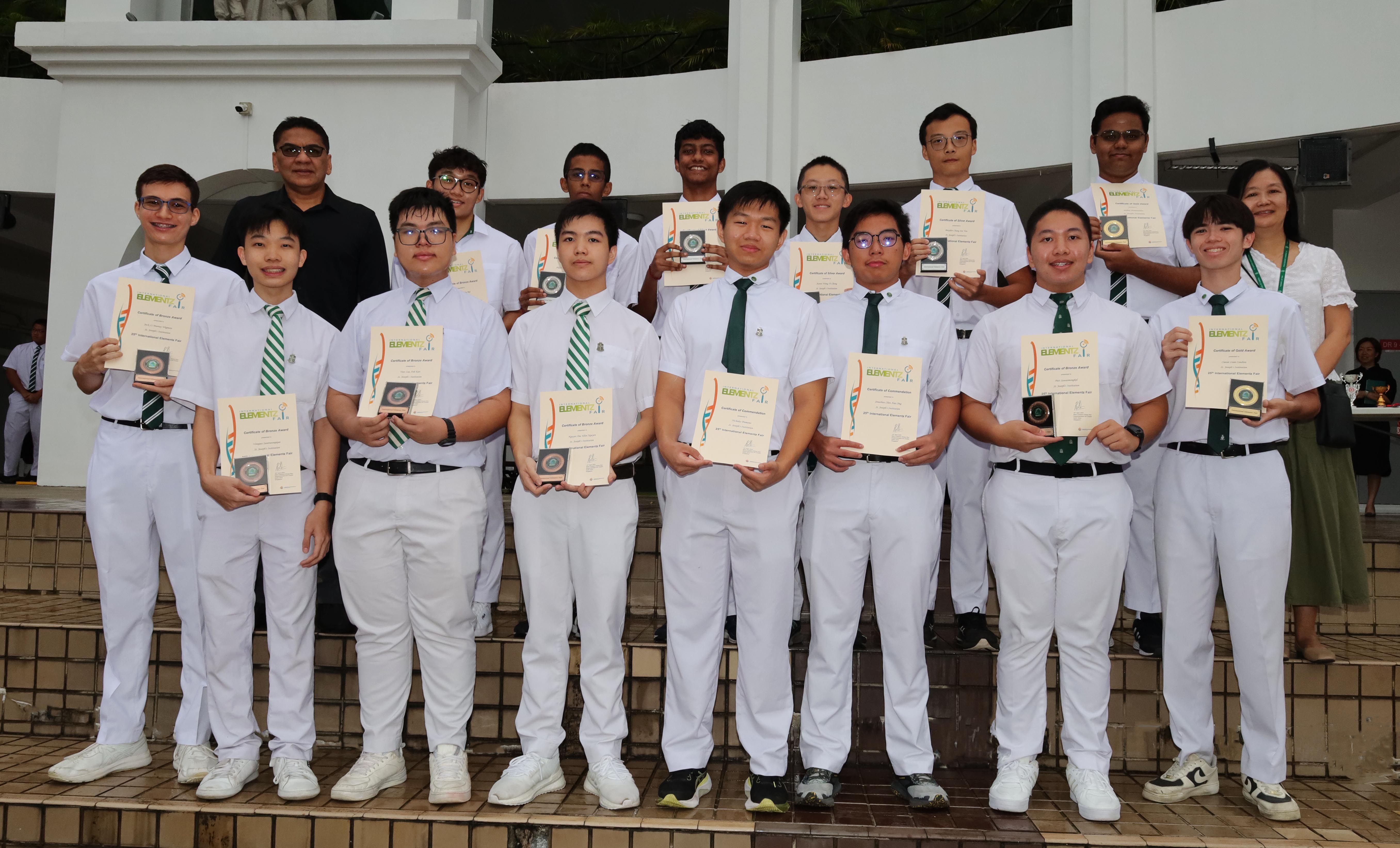 A group of students in white uniforms holding certificates and trophies, posing outdoors with two adults.